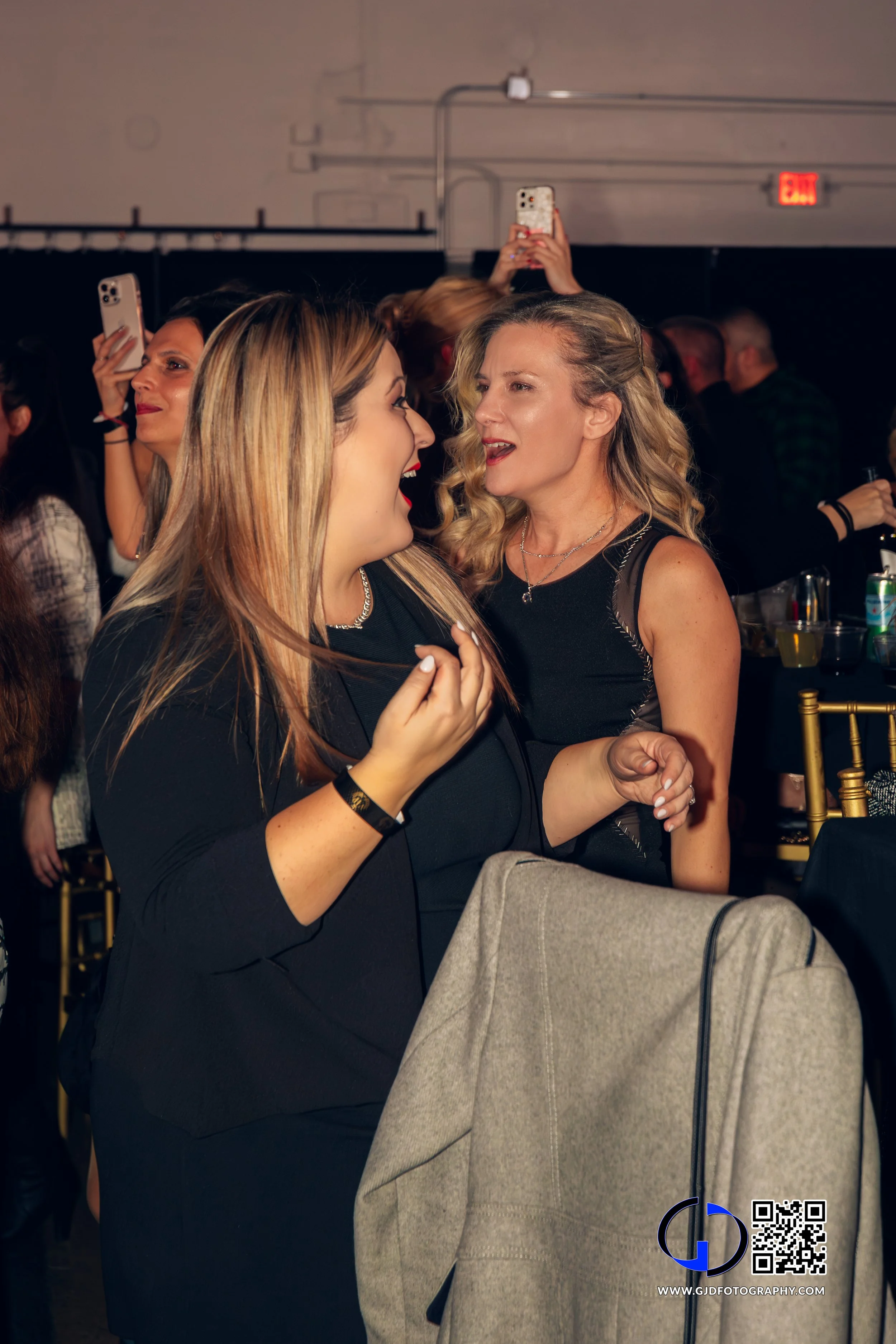 Two women in black outfits are talking and laughing at a crowded indoor event, with several people behind them taking photos with their phones.