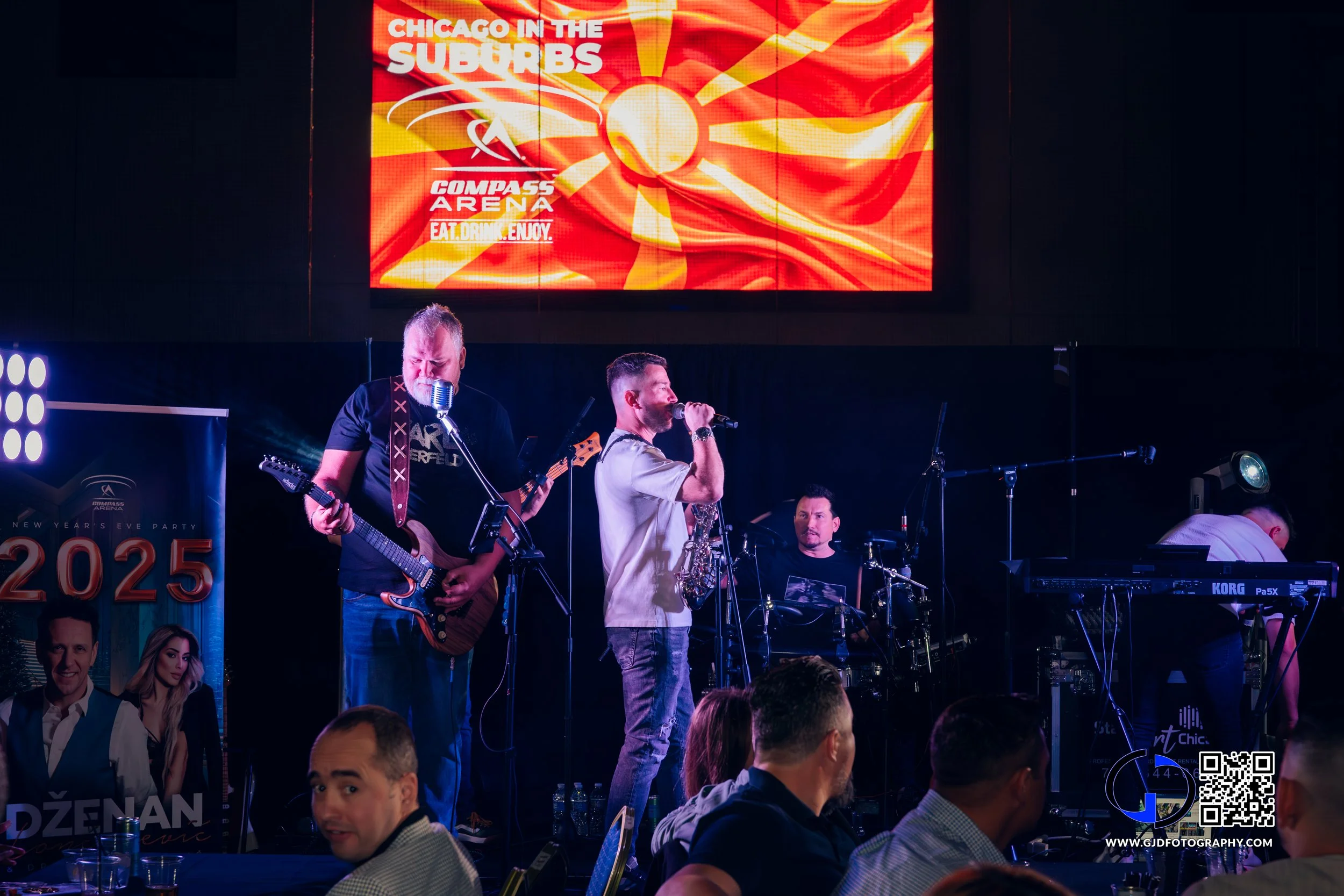 A live music performance on stage featuring three musicians, one playing guitar, one singing into a microphone, and one on drums, with an audience in the foreground and a large screen above displaying a graphic promoting an event at Compass Arena.
