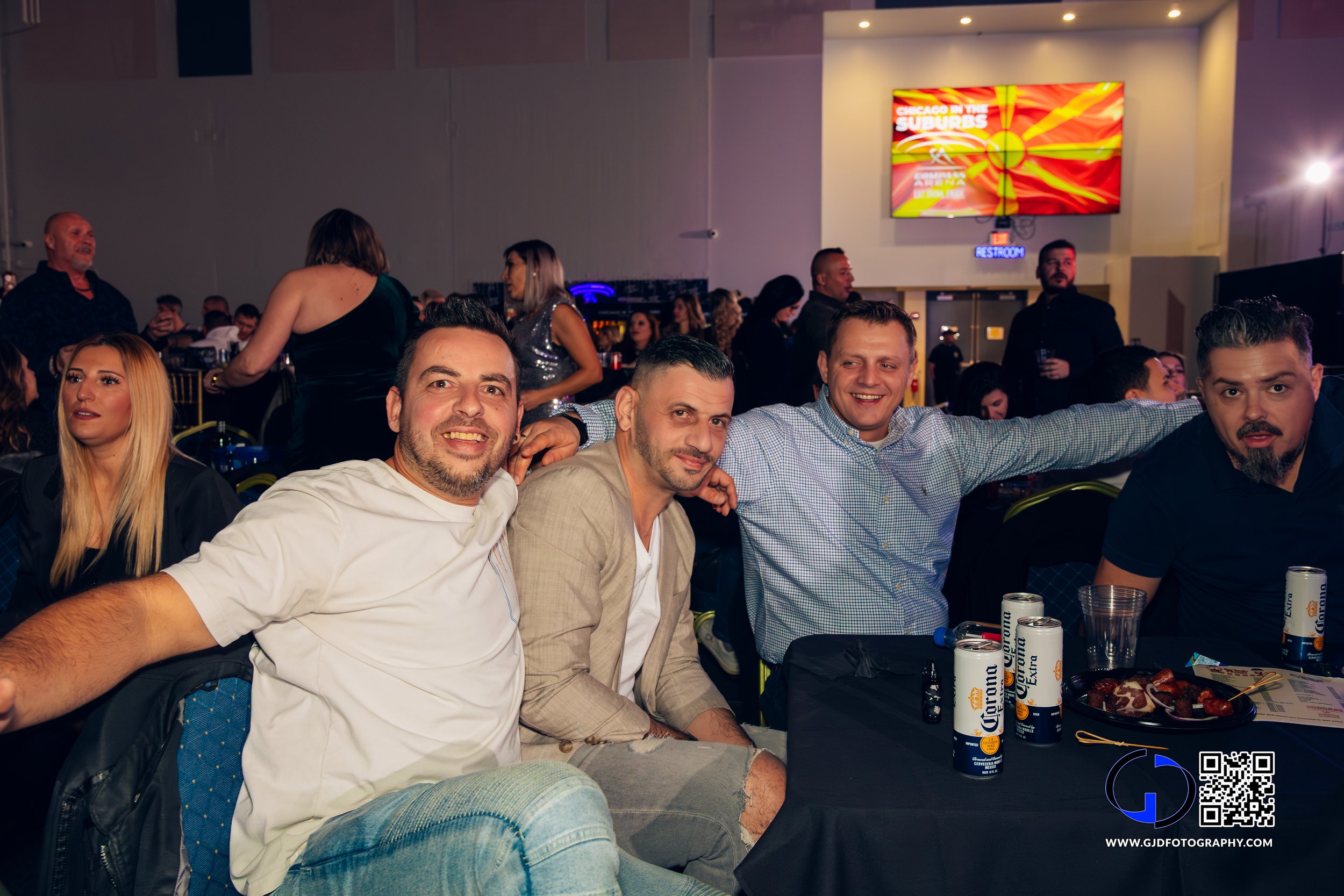 Group of four men sitting at a table with drinks and food, smiling at a lively event or party in a large hall with a crowd in the background.