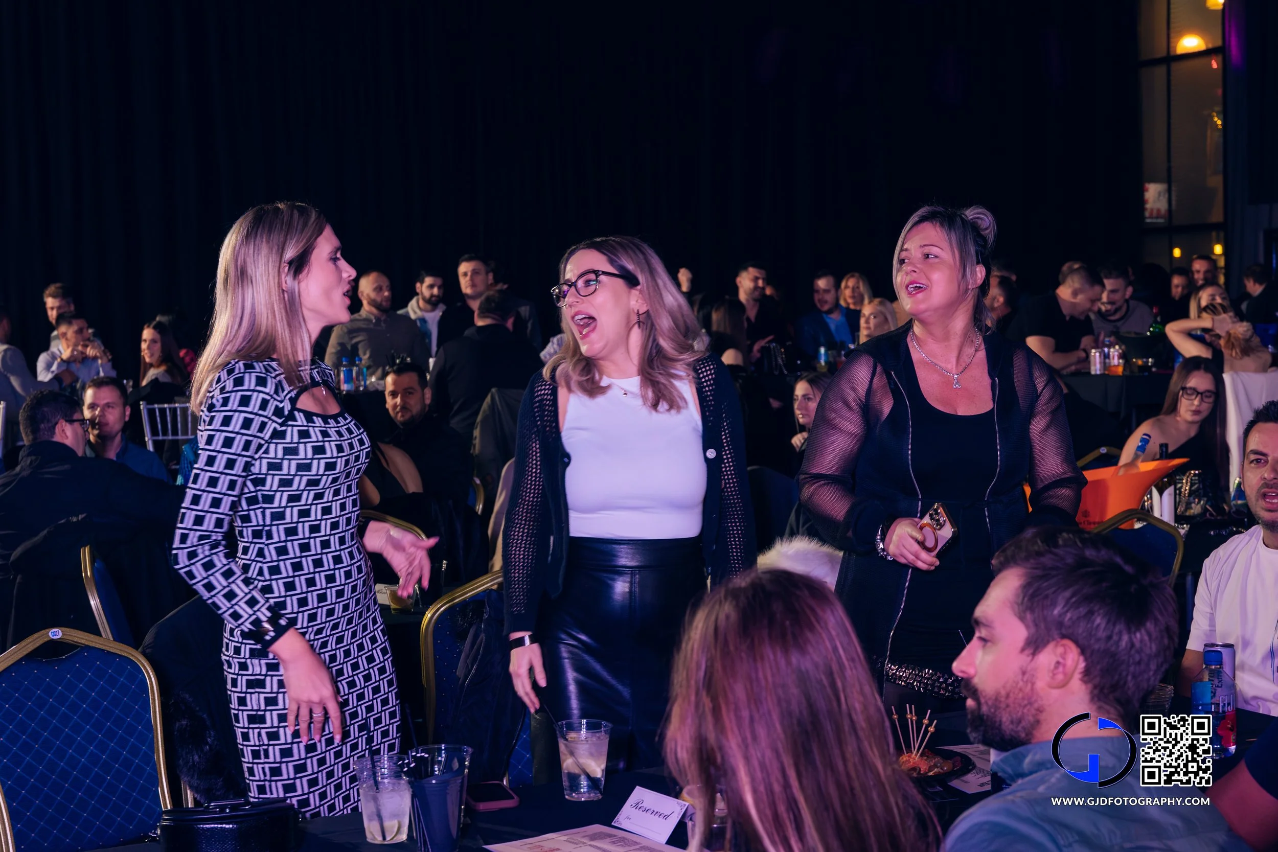 Three women are standing and talking at a crowded event with many seated guests in the background.