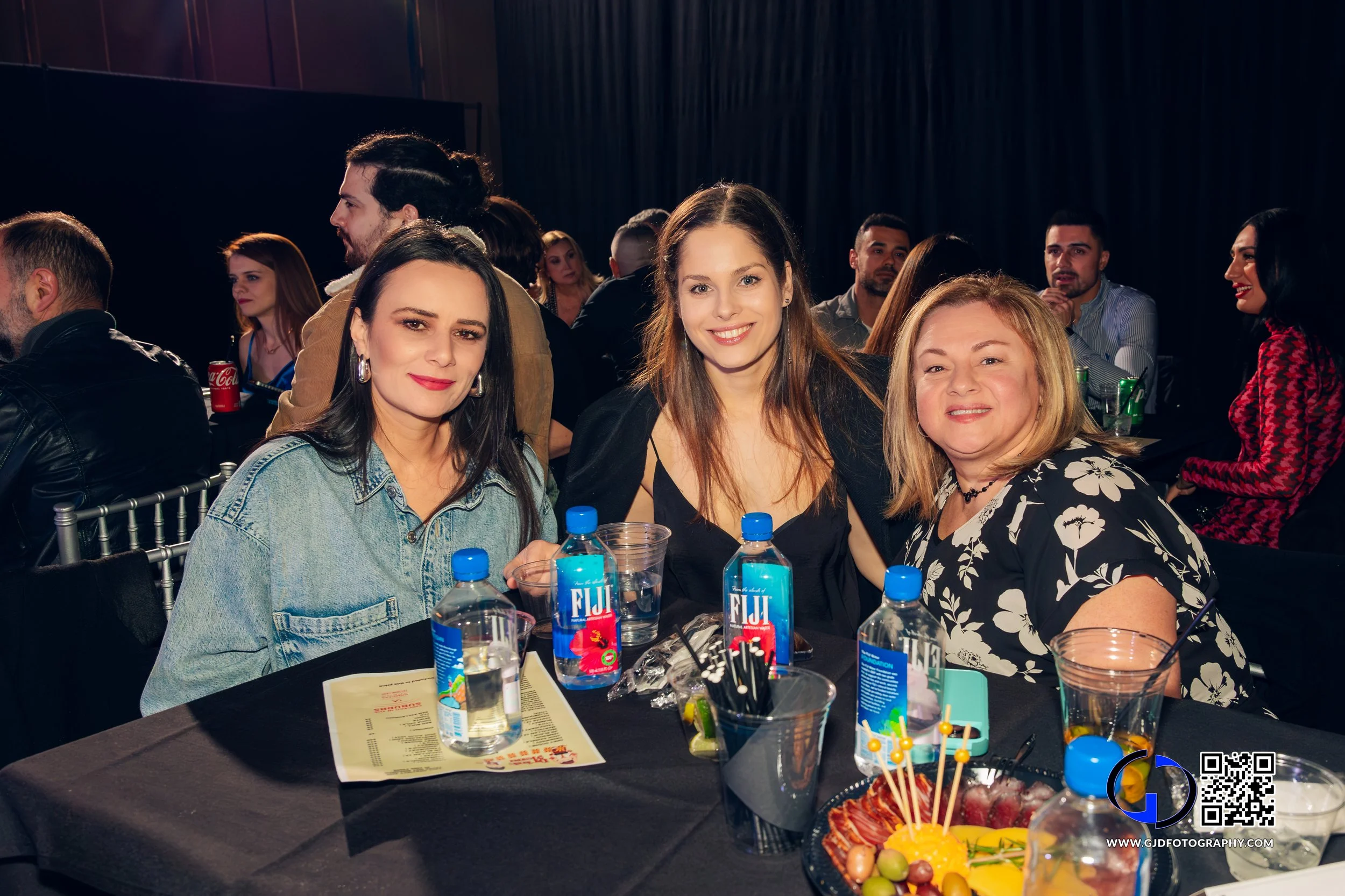 Three women sitting at a table with drinks, bottles of Fiji water, a platter of fruit, and party decorations at an indoor event.