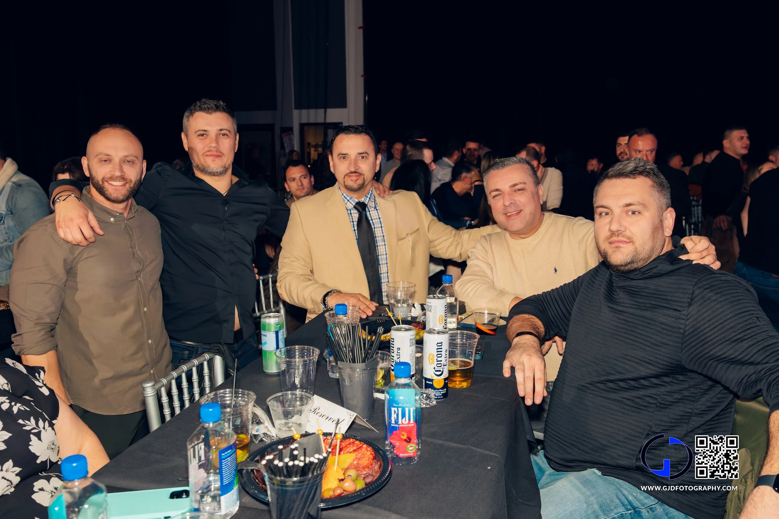 Group of five men gathered around a table at a social event, with drinks and snacks, smiling and posing for the camera.