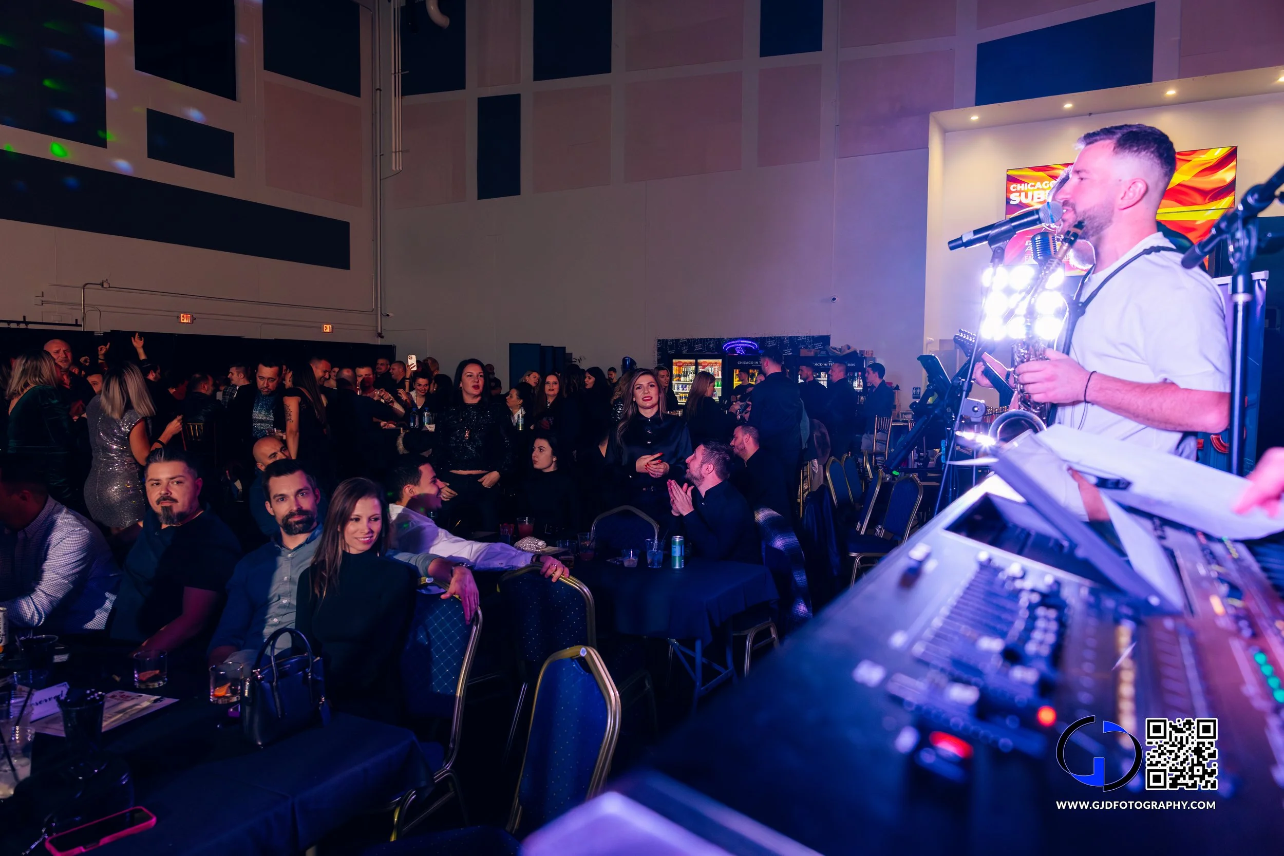 A man performing music or singing on stage with a microphone, while an audience of mostly young adults watches in a dimly lit, indoor event space. The crowd is seated at tables and standing, with some talking and drinking. There is a DJ or sound equi