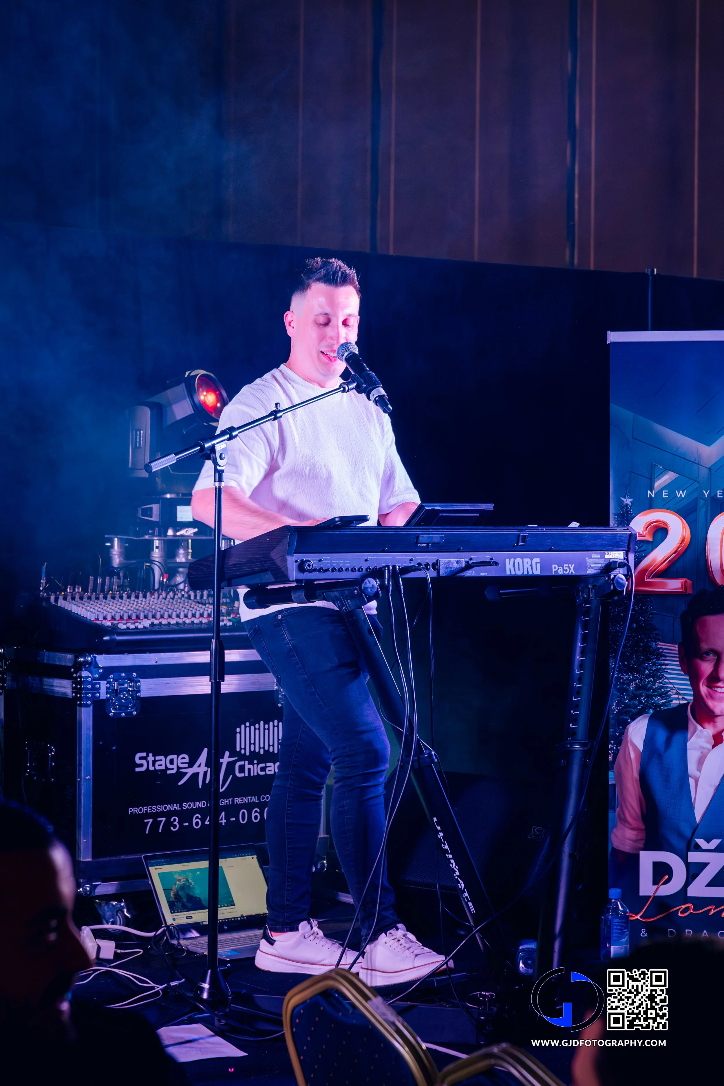 A male musician performing on stage with a keyboard and microphone, wearing a white shirt and jeans, under stage lights.