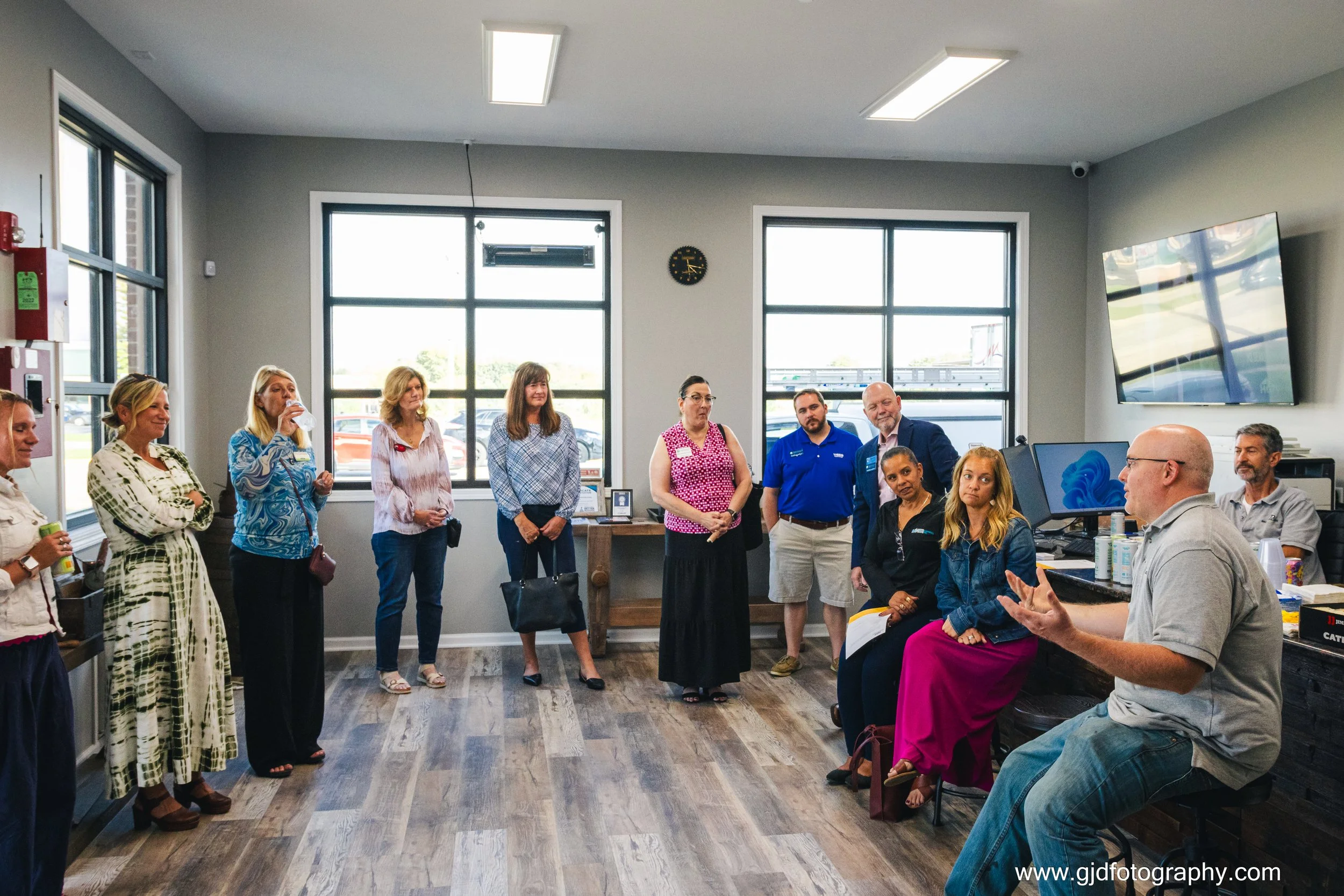 A group of people standing and sitting in a modern office or meeting room with large windows, listening to a man in a gray shirt who is speaking and gesturing with his hands.