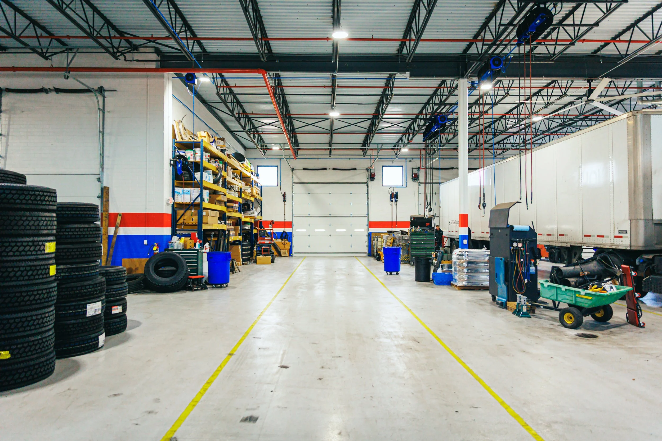 A large, well-lit truck repair garage with tires stacked on the left, shelves with tools and supplies along the back and right, and a white truck trailer on the right side. The garage has a concrete floor with yellow lines and a high ceiling with exp