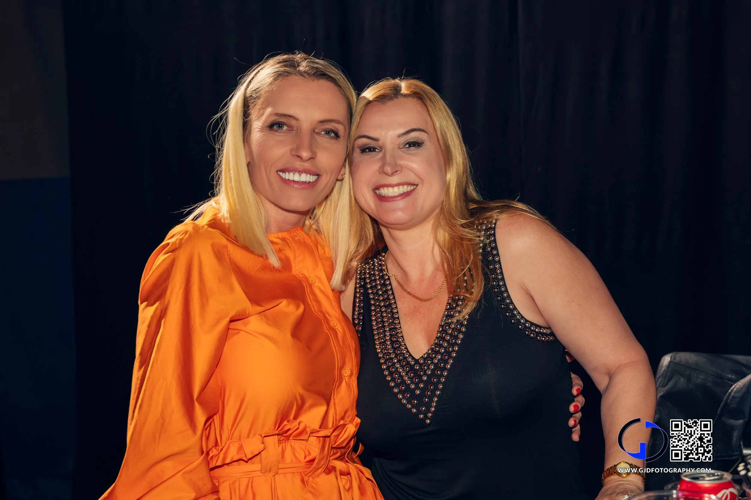 Two women smiling and posing together indoors, one wearing an orange dress and the other a black sleeveless top with embellished neckline.