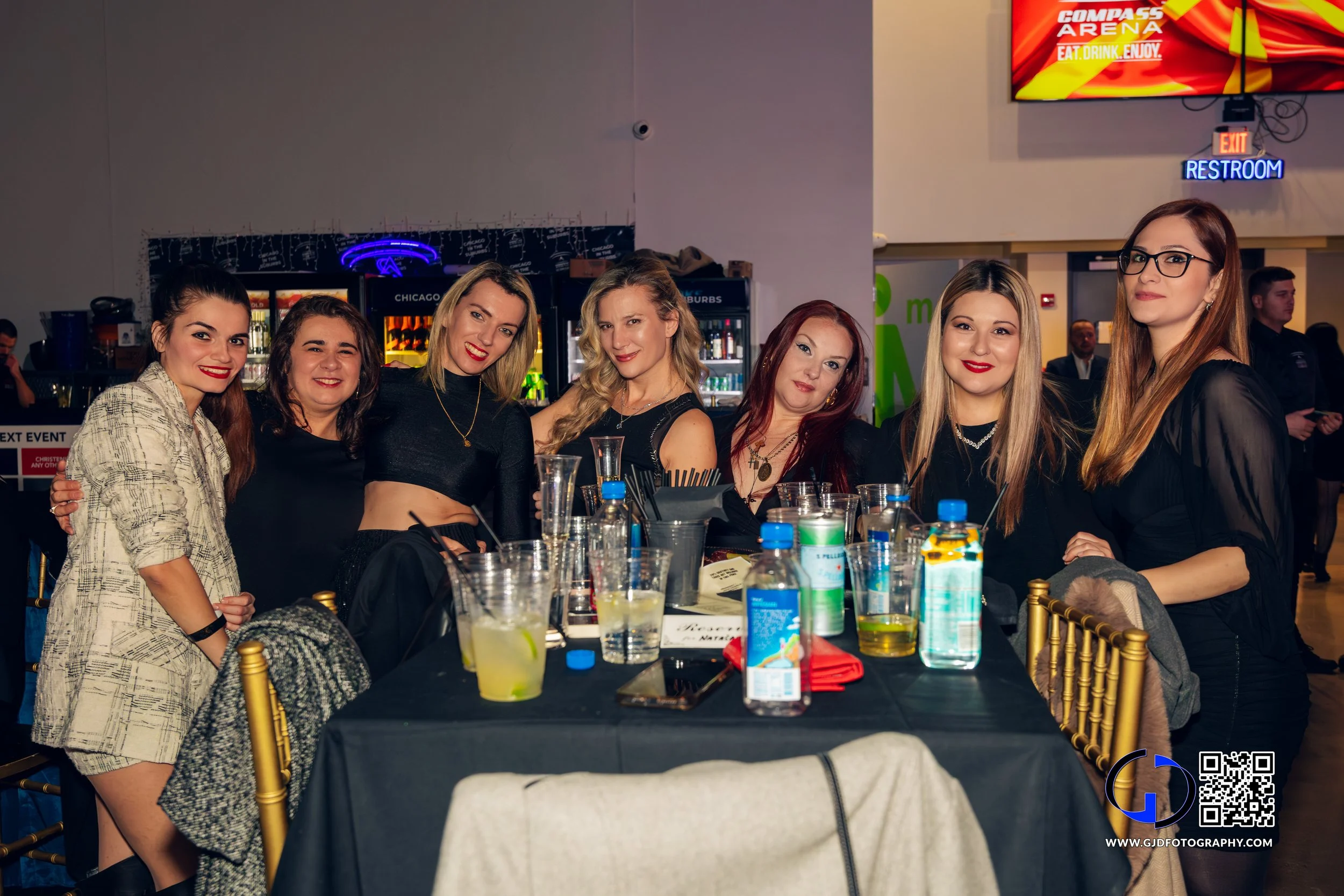 Group of eight women seated at a table in a lively indoor venue, smiling and posing for the photo, with drinks and personal items on the table. Behind them is a bar area and a digital screen displaying bright red and yellow graphics.