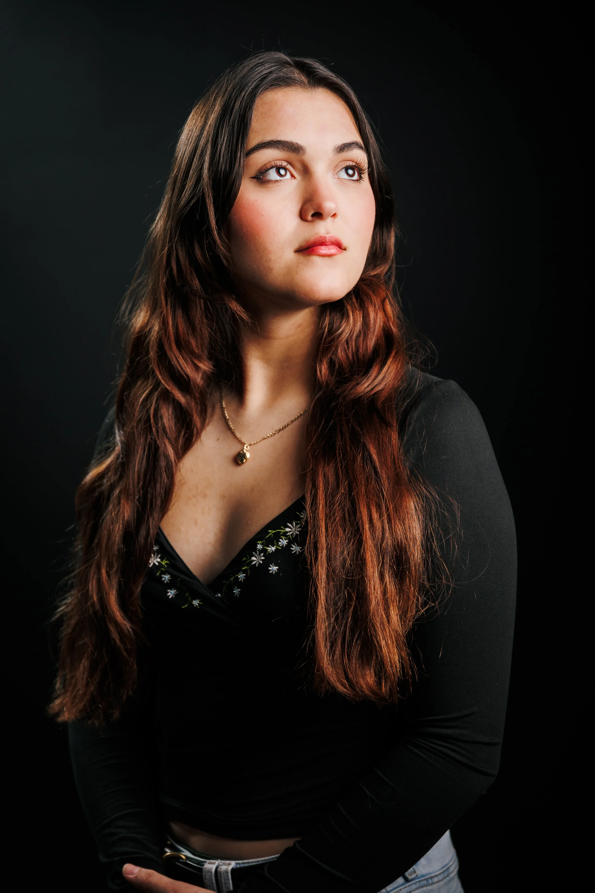 A young woman with long, wavy brown and red hair, wearing a black top with floral embroidery, a gold necklace, and jeans, looking slightly upward against a dark background.