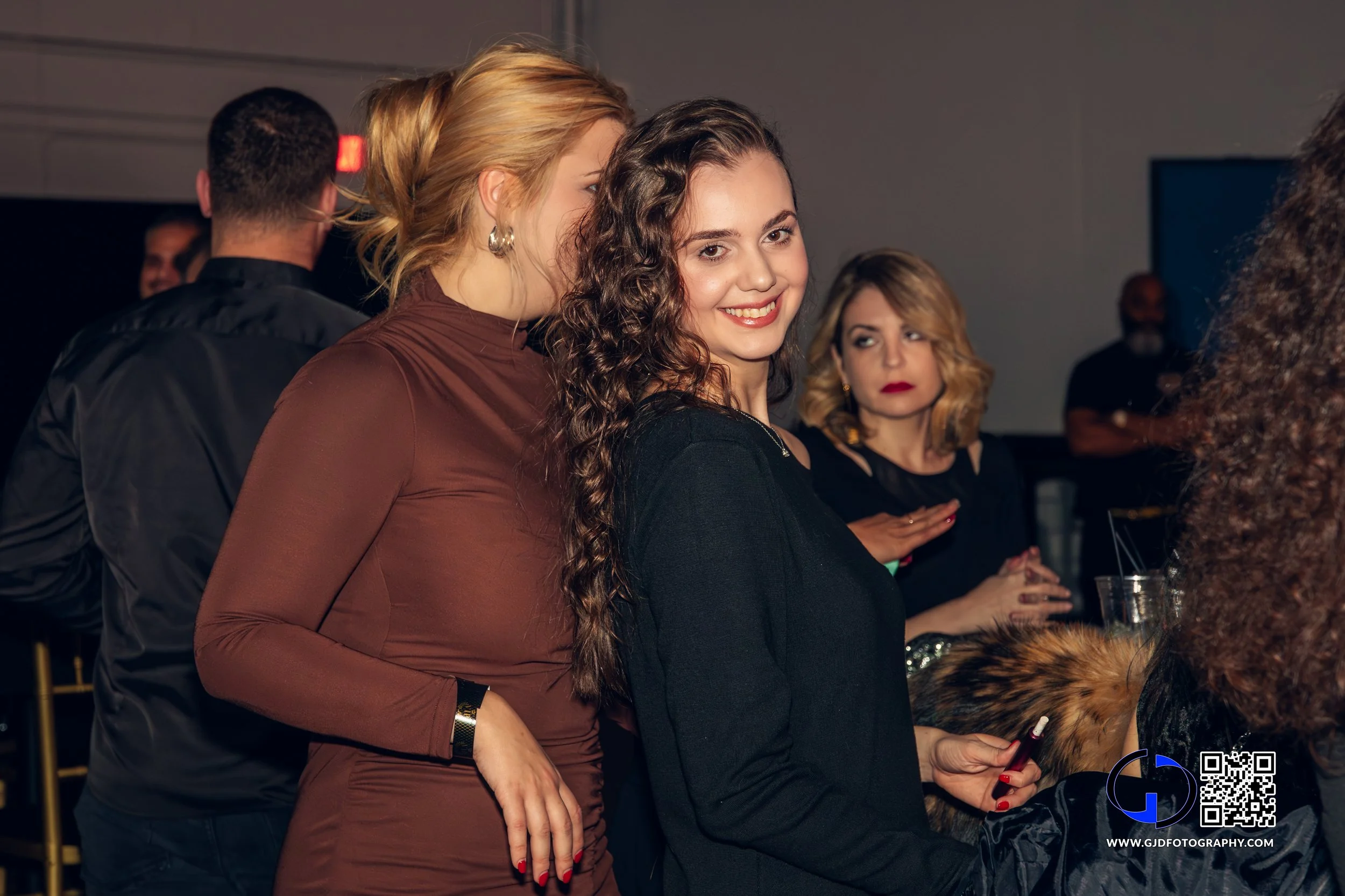 A woman with long curly brown hair smiling at an event, wearing a black top. She is surrounded by other people, including a woman with blonde hair and a woman with blond hair and red lipstick.