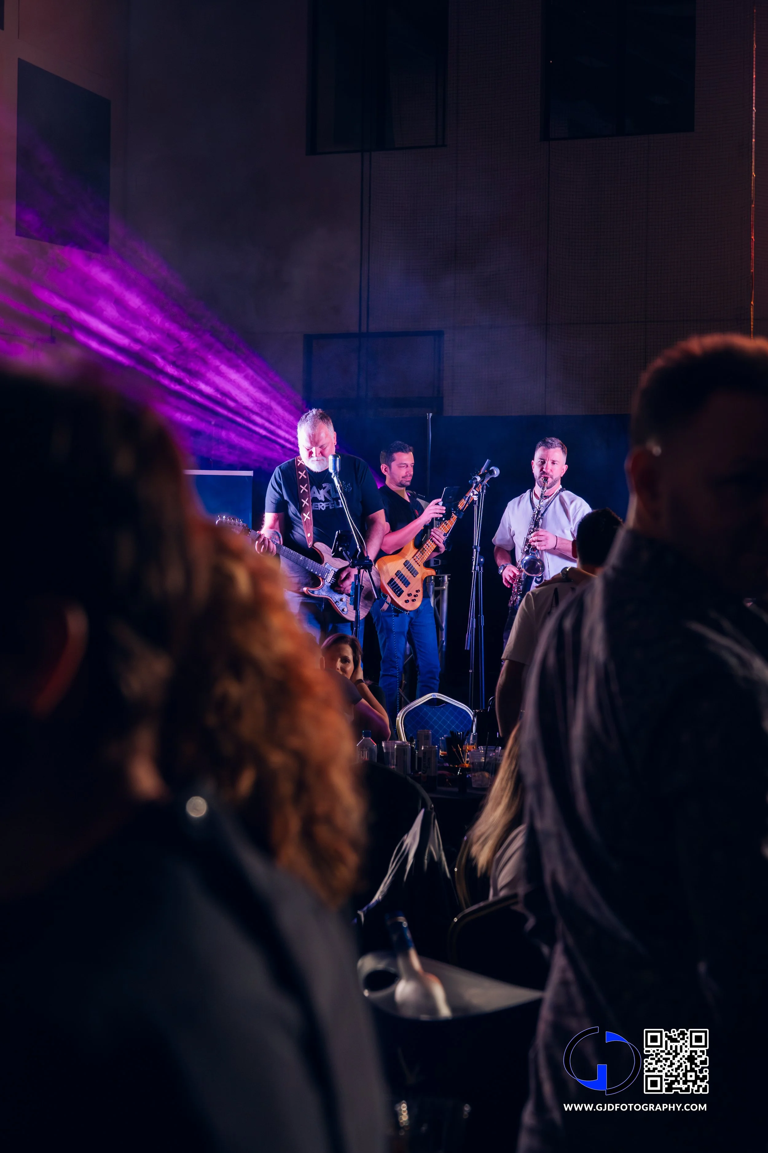 Live band performing on stage with purple lighting, audience seated at tables in a dark venue.