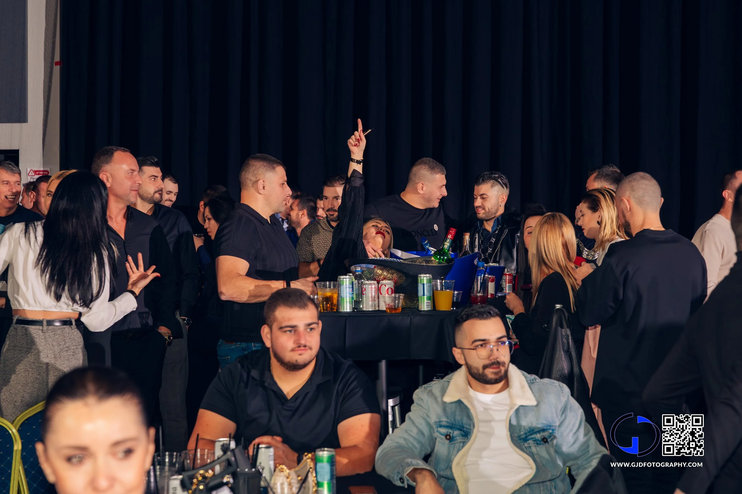 People socializing at a party around a table with drinks, with one woman in black raising her hand and leaning on the table, against a black curtain backdrop.