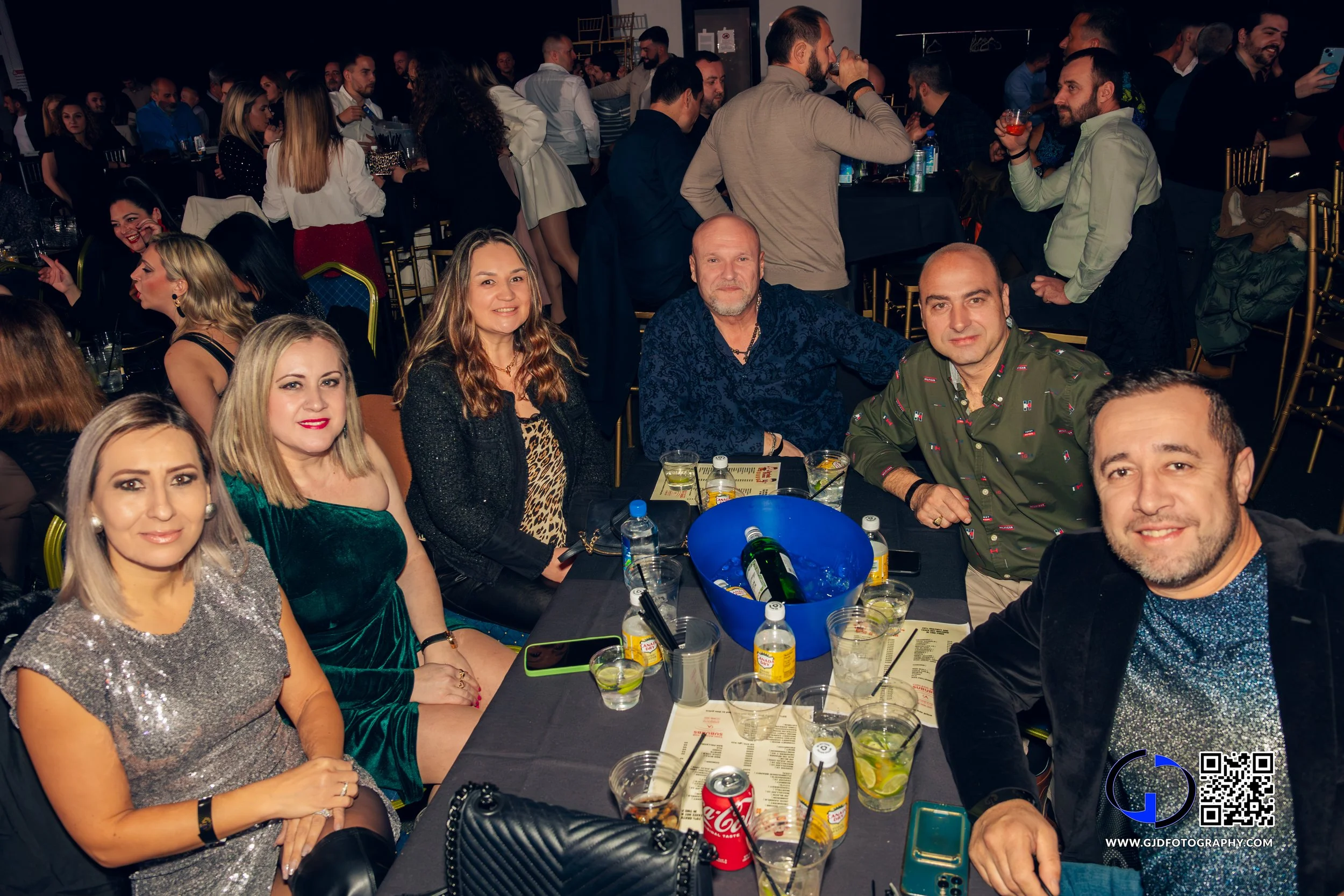 Six people sitting at a table during a celebration event, with drinks and snacks, in a crowded venue with other groups in the background.