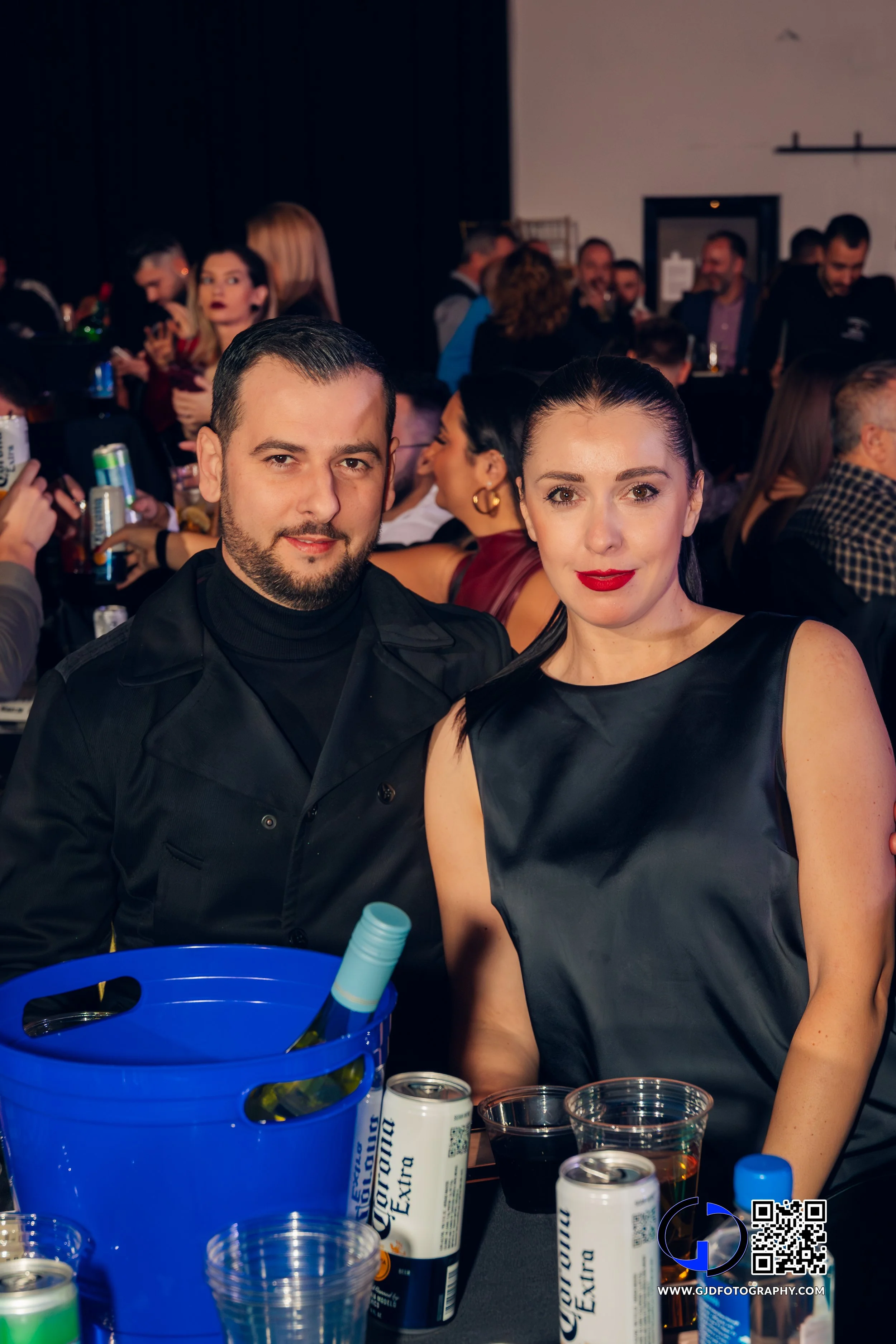 A man and a woman sitting together at a social event or party with drinks and cans on the table, surrounded by many people in the background.