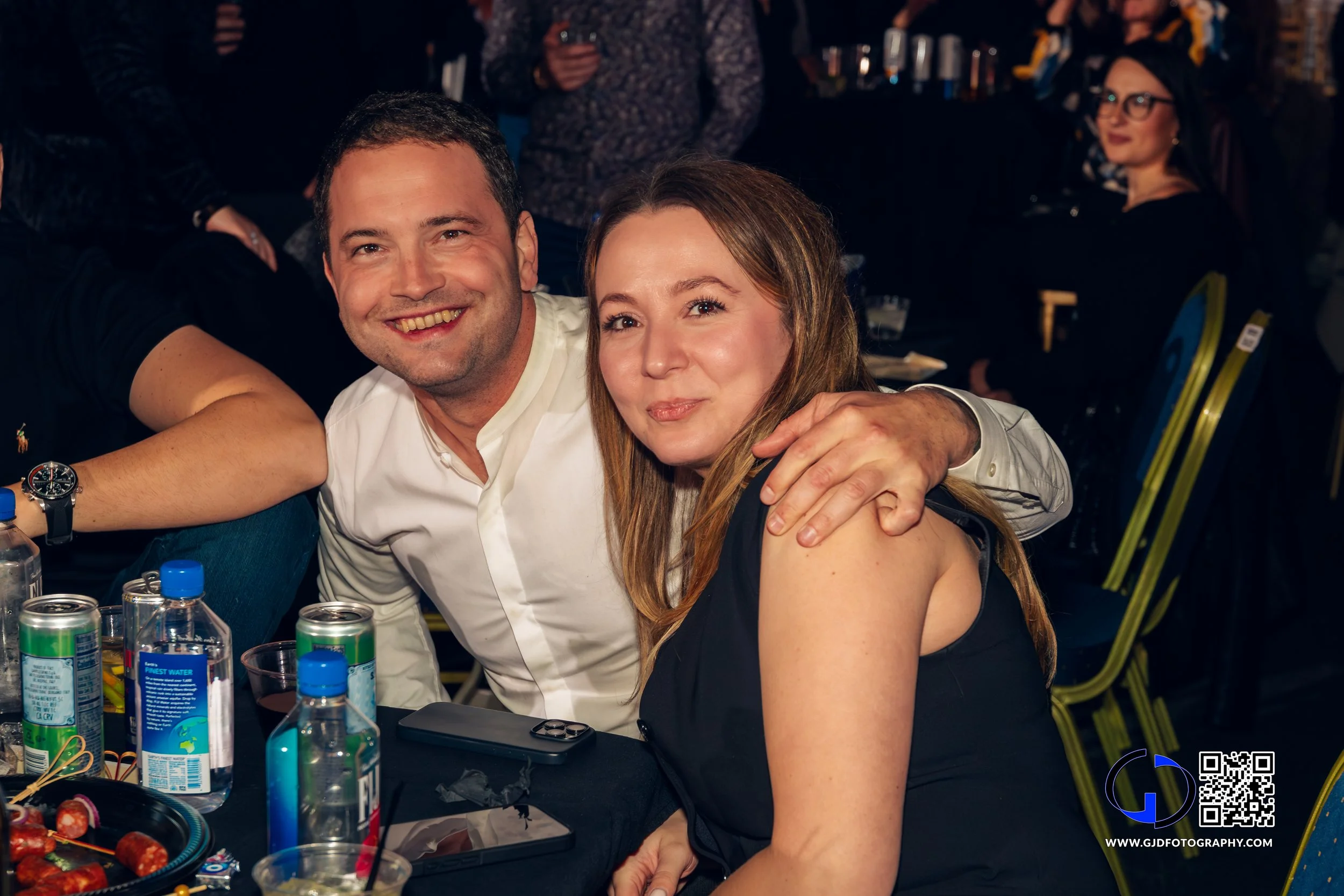 A man and woman smiling at a social gathering, with drinks and snacks on the table in front of them.