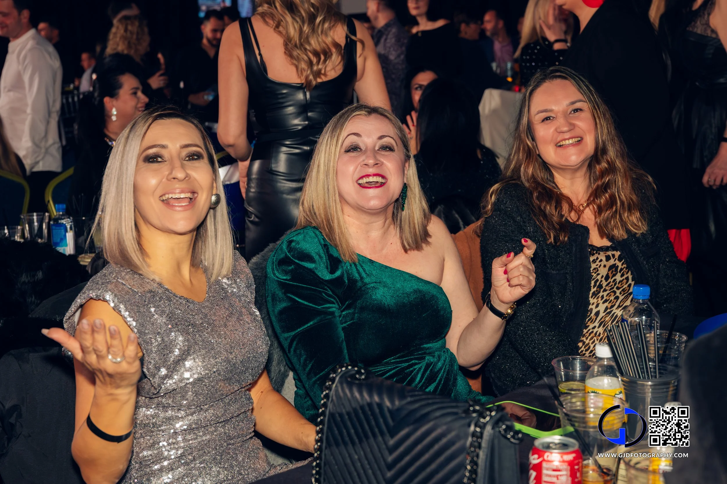Three women smiling and posing at a social event, sitting at a table with drinks and snacks, surrounded by other people in a lively setting.