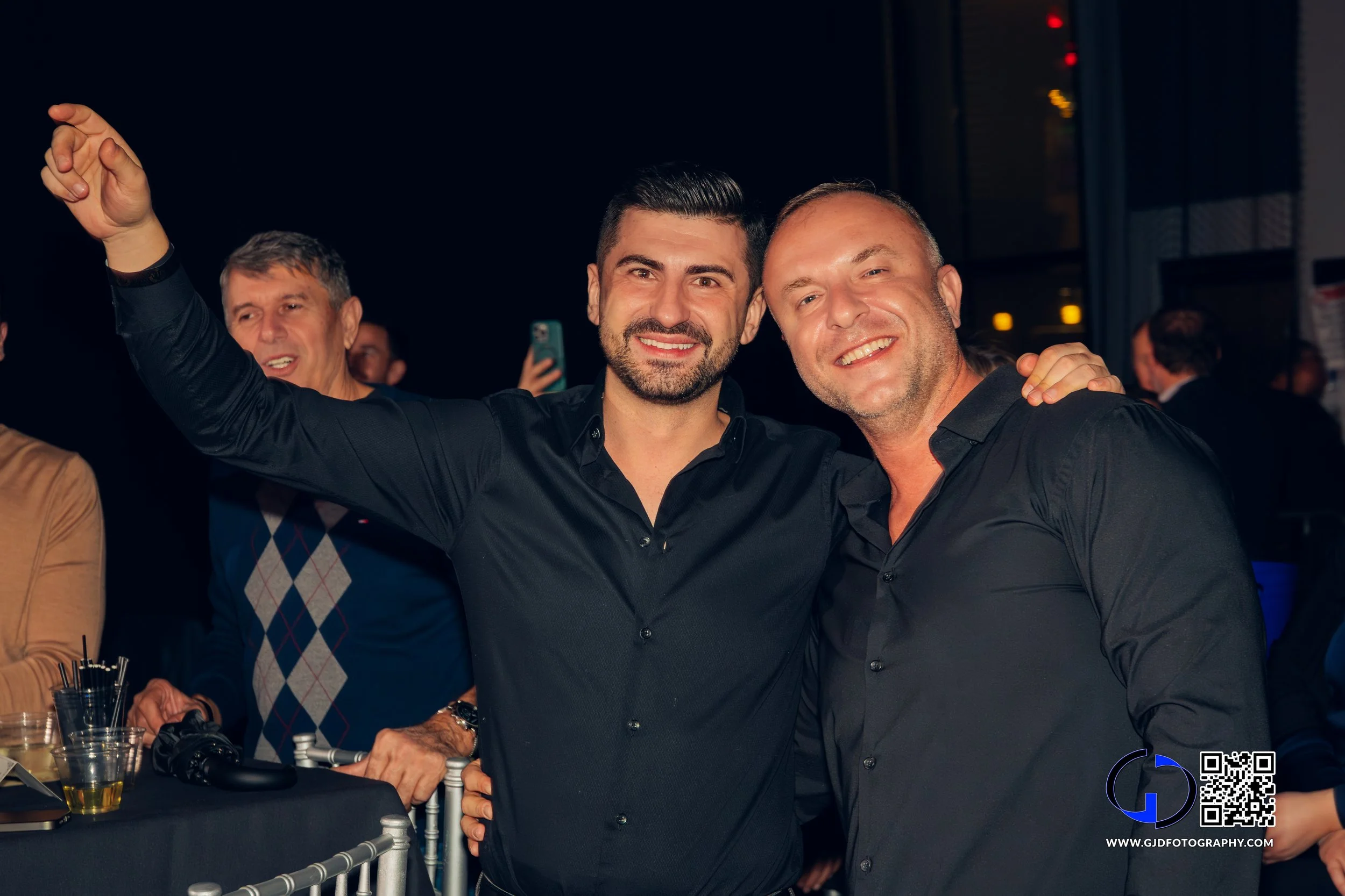 Two men smiling and posing together at a social event with one man's arm around the other's shoulder, in a dimly lit room with other people in the background.