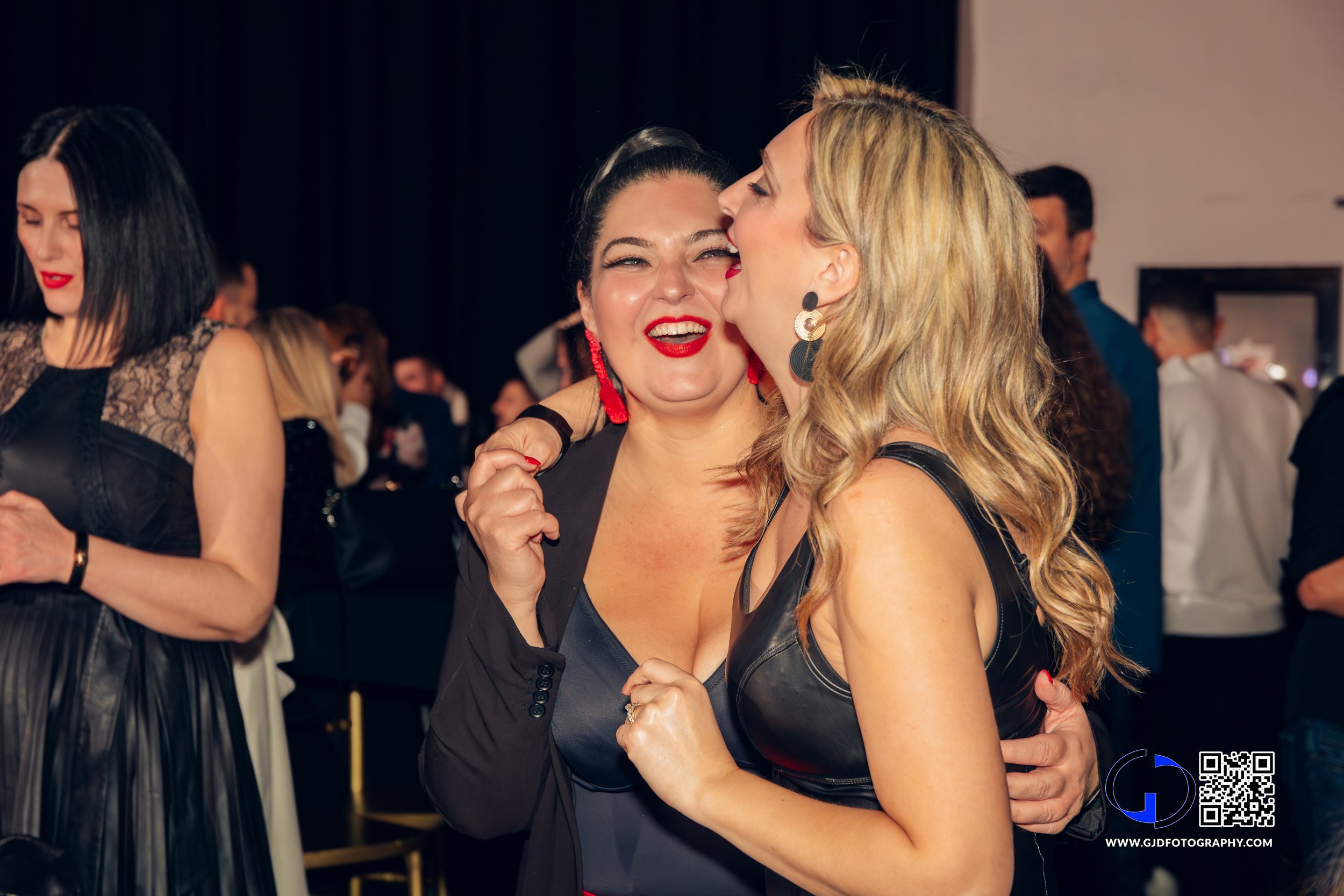 Two women dancing and laughing at a party, one whispering to the other, with others in the background.