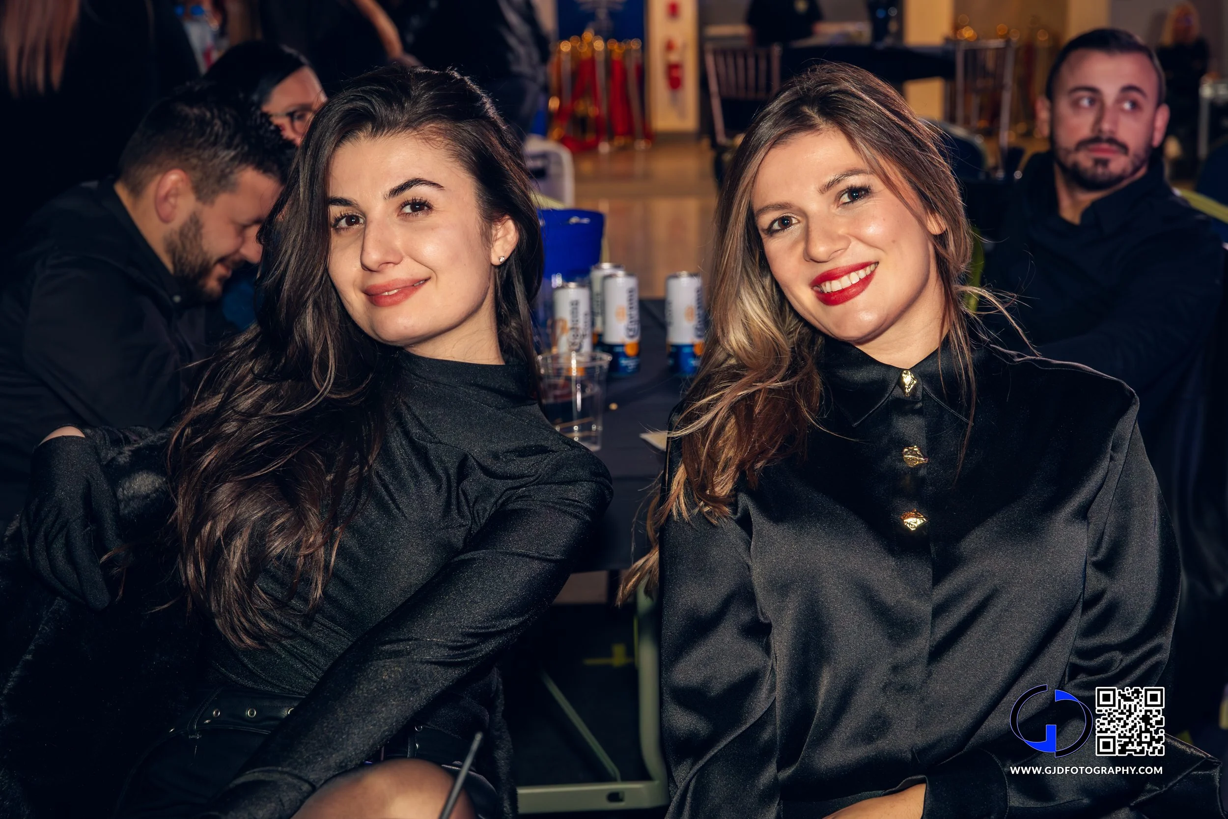 Two young women sitting at a table, smiling at the camera, with a blurred background of people and event decorations.