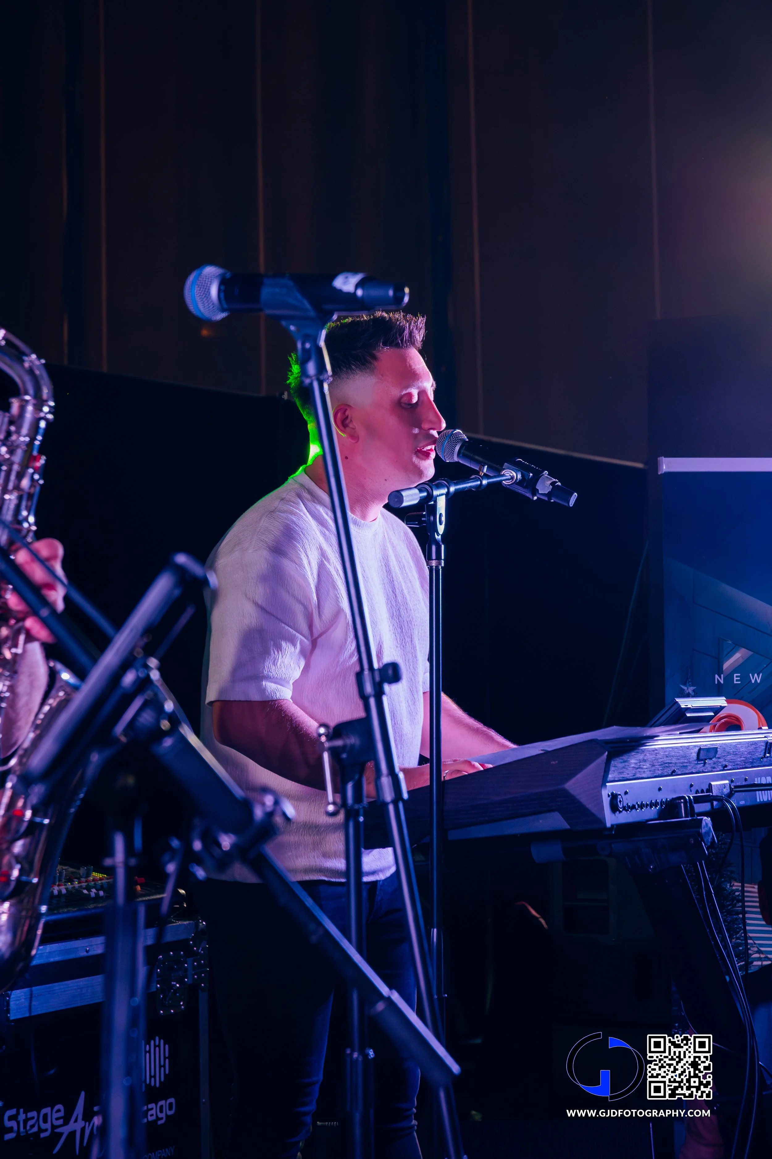 A musician with a mohawk hairstyle singing into a microphone while playing a keyboard on stage, with colorful stage lights.