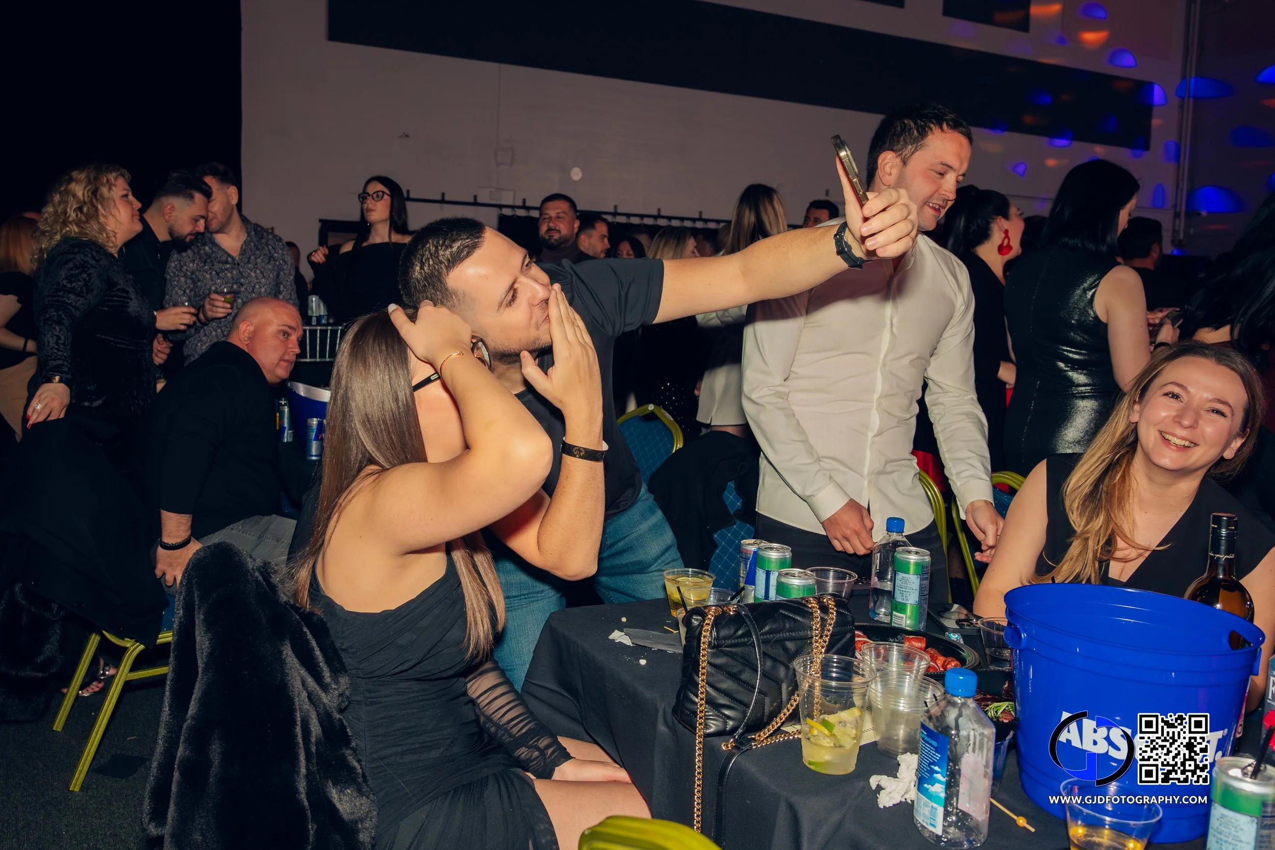 People at a party, taking a selfie, some smiling and some covering their ears, with drinks and bottles on the table.