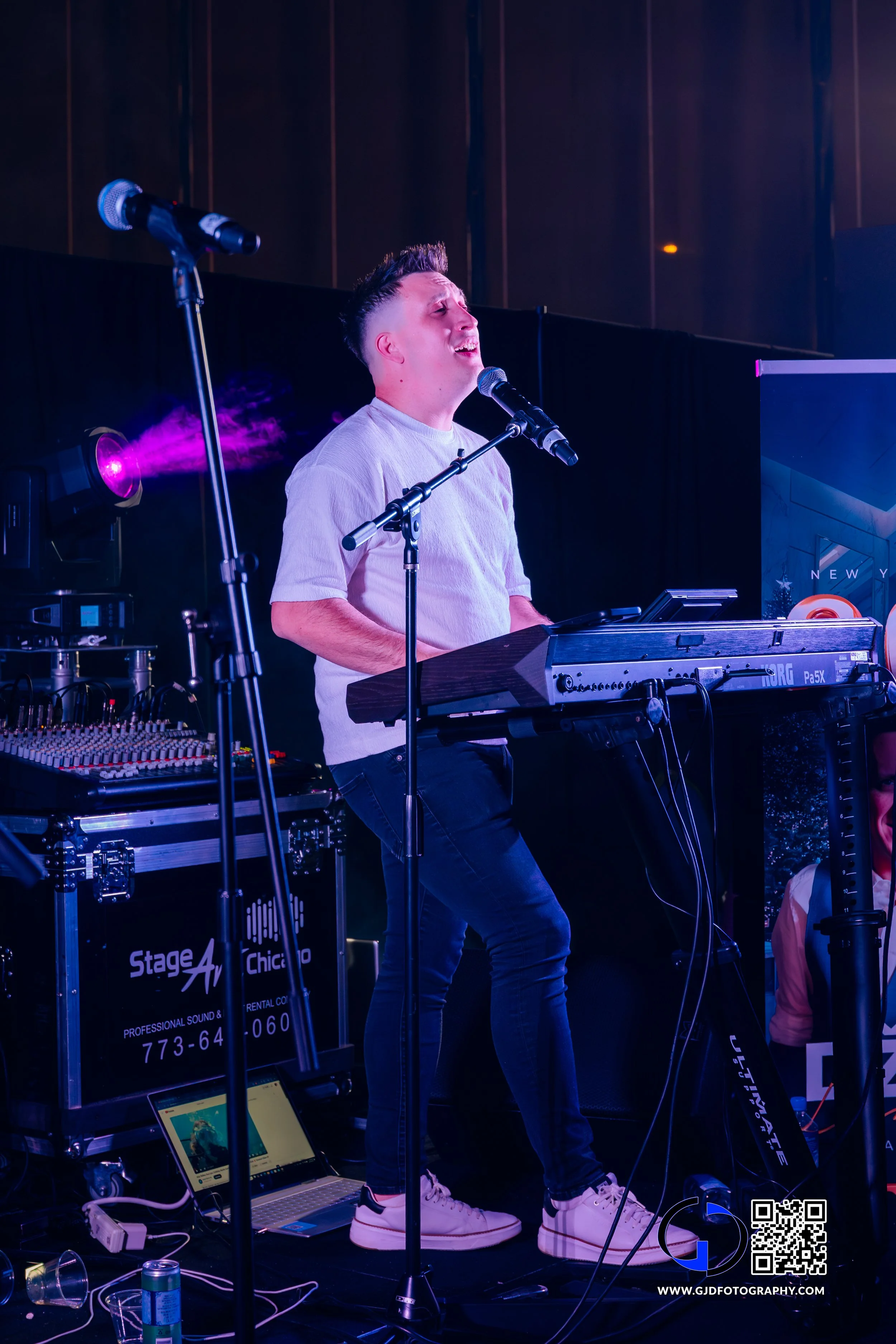 A person performing live on stage singing with a keyboard, microphone, and DJ equipment, with purple stage lighting.