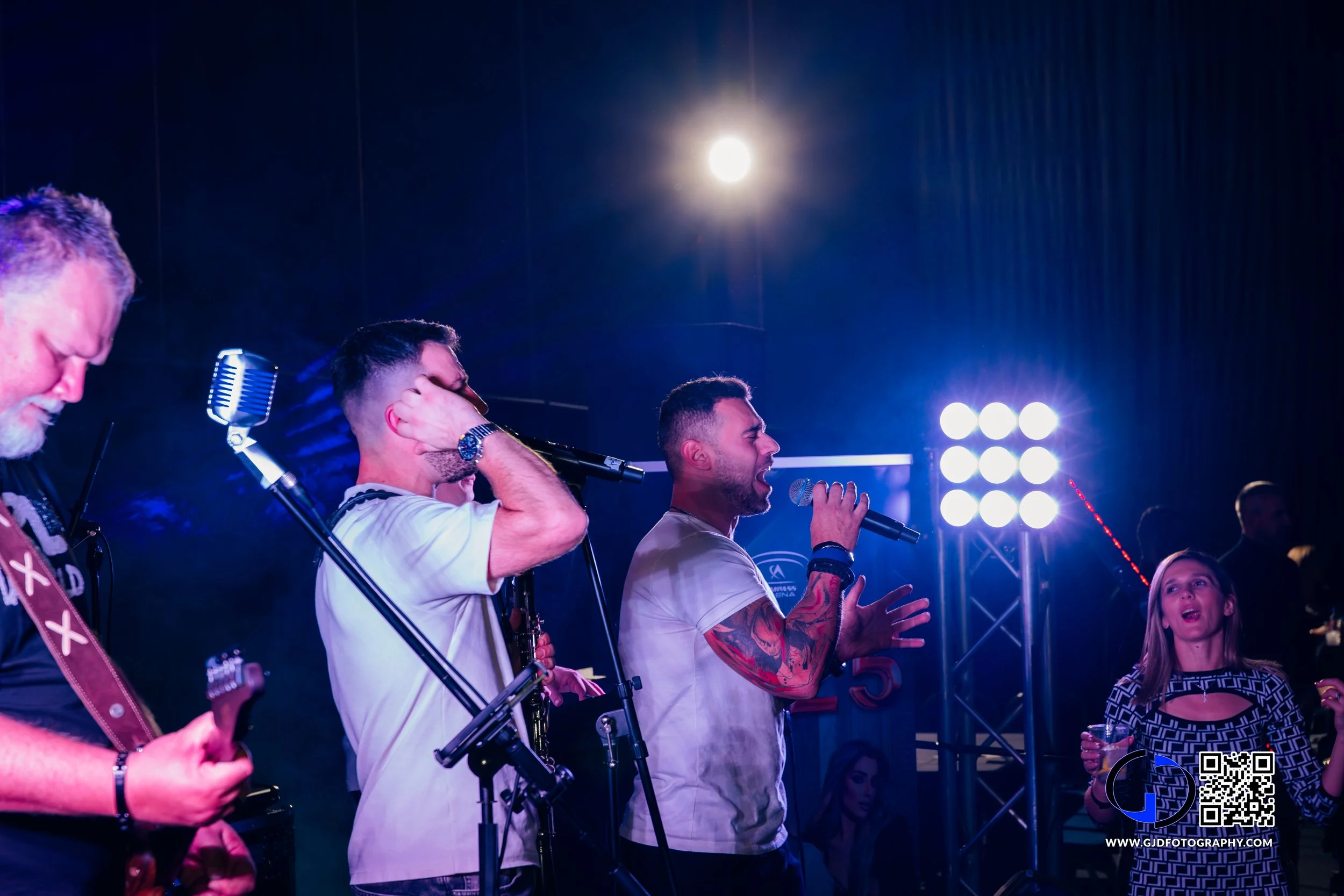 Music band performing on stage with female singer holding microphone, band members playing guitar and instrument, stage lighting, and audience in background.