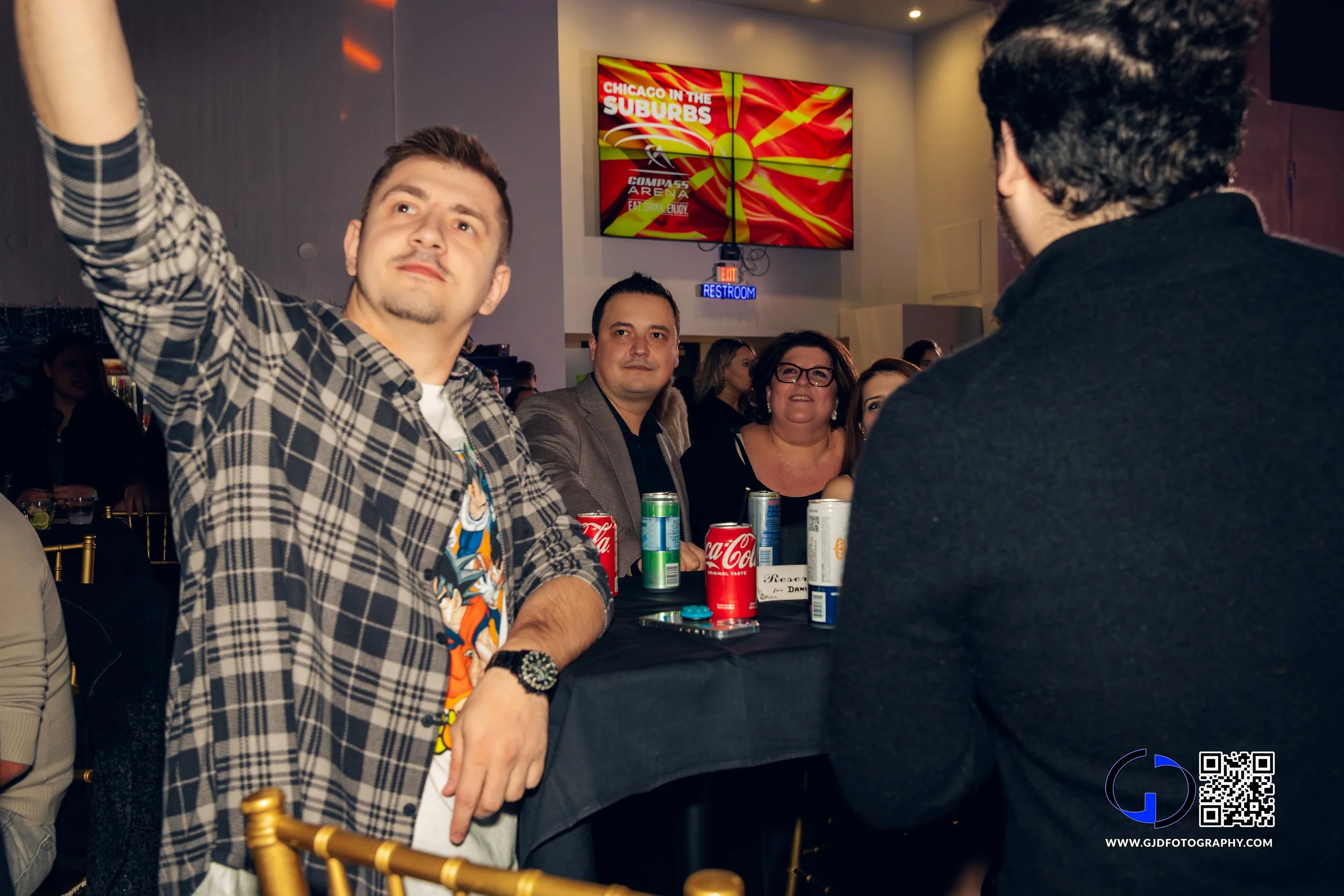 Group of people sitting at a table in a dimly lit event space, some looking towards the camera, with drinks and cans of Coke on the table, and a digital screen showing 'Chicago in the Suburbs' in the background.