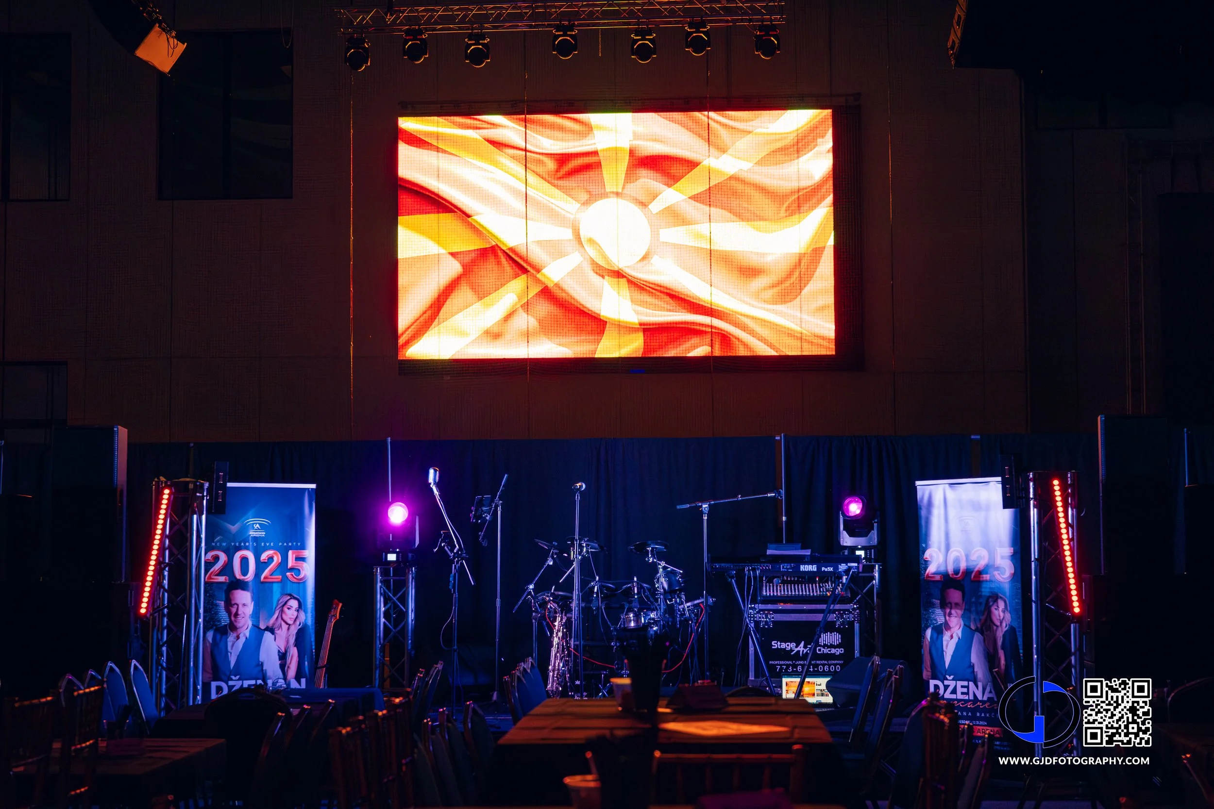 An indoor stage set up for a New Year's Eve event, with musical instruments, banners displaying '2025' and images of a man and woman, and a large LED screen showing an abstract fiery sun. There are purple and red stage lights and chairs arranged in f