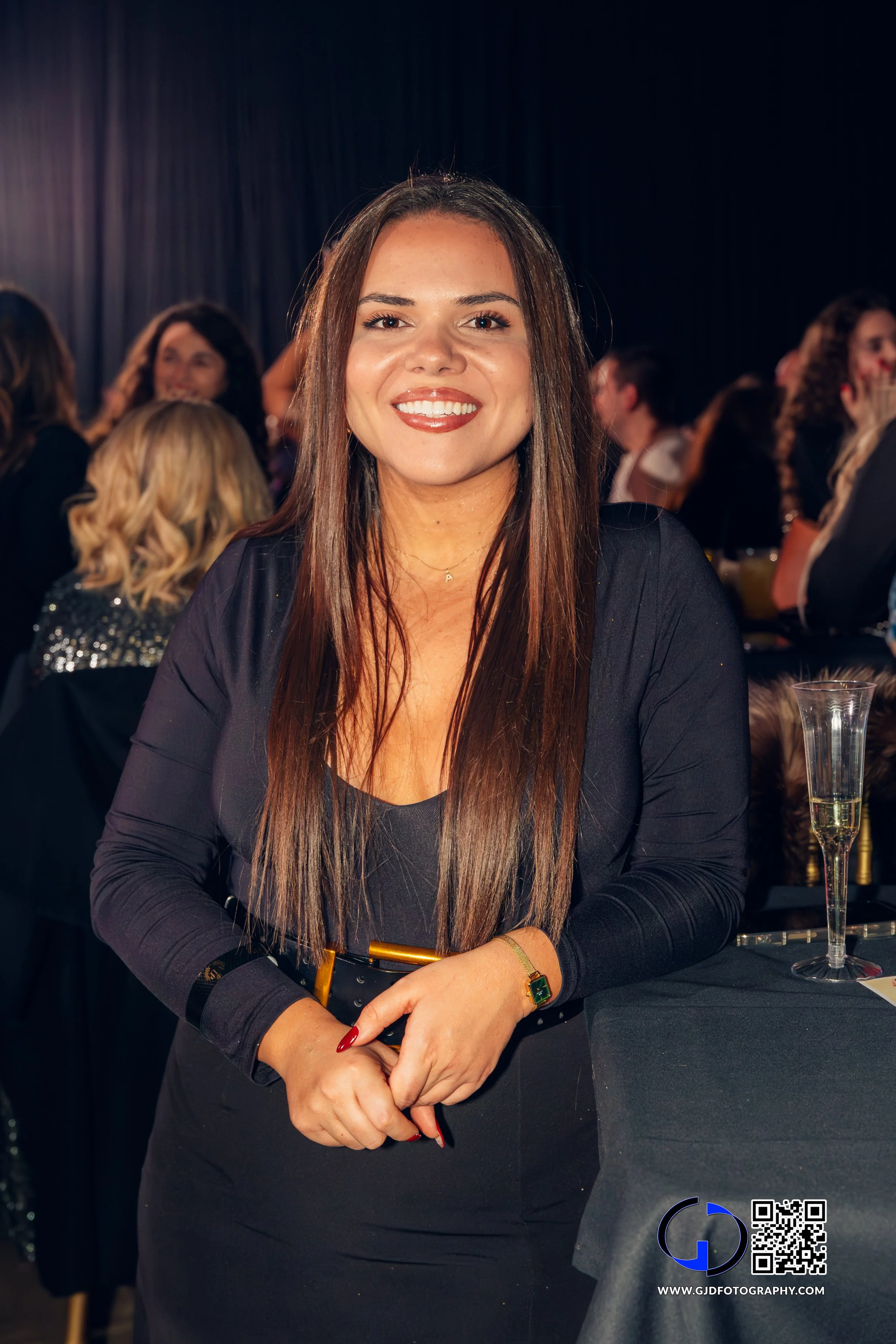 A woman with long brown hair and a black dress smiles at a social event with people in the background and a glass of champagne on the table.