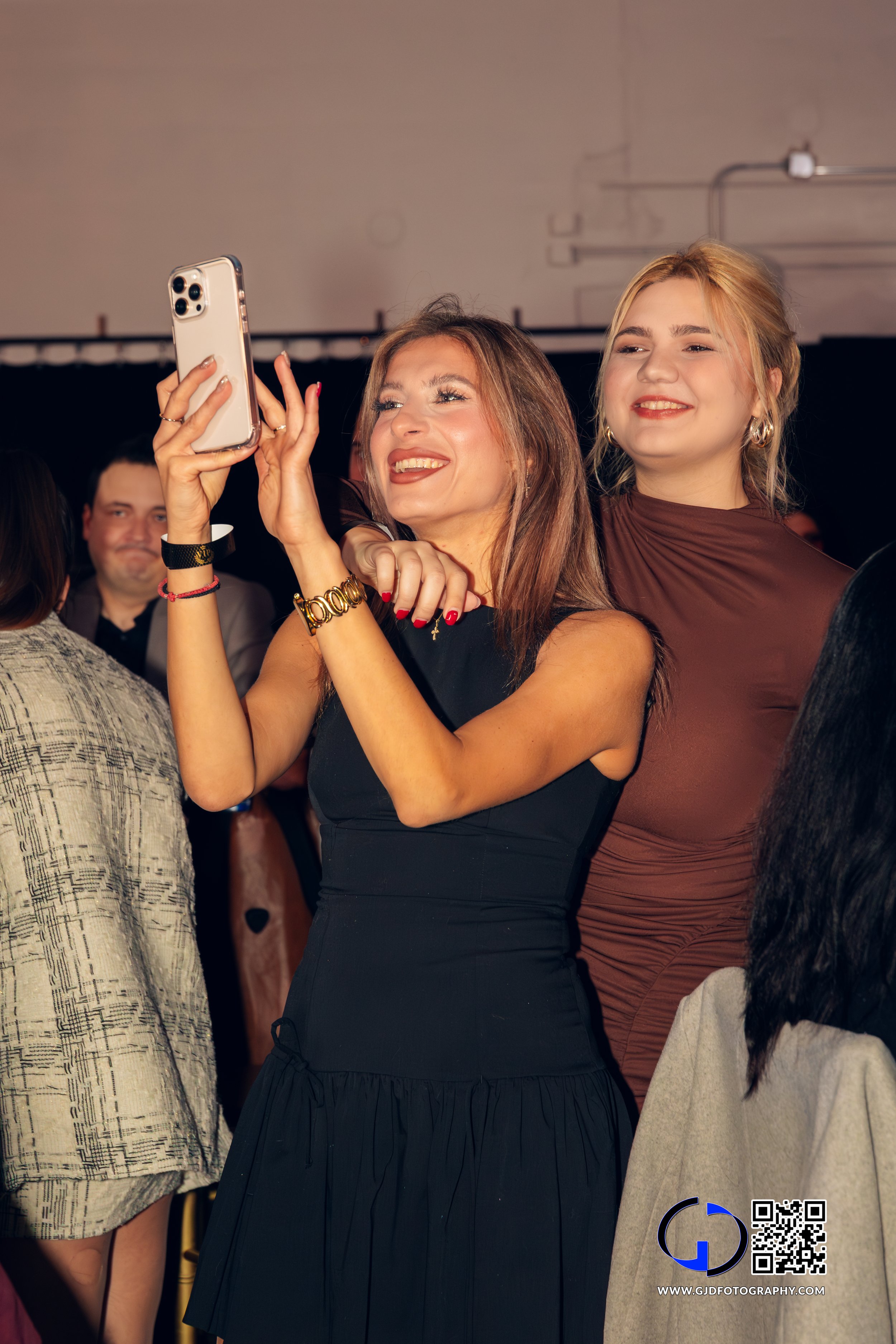 Two women taking a selfie at a social event, with smiley faces and a crowd in the background.