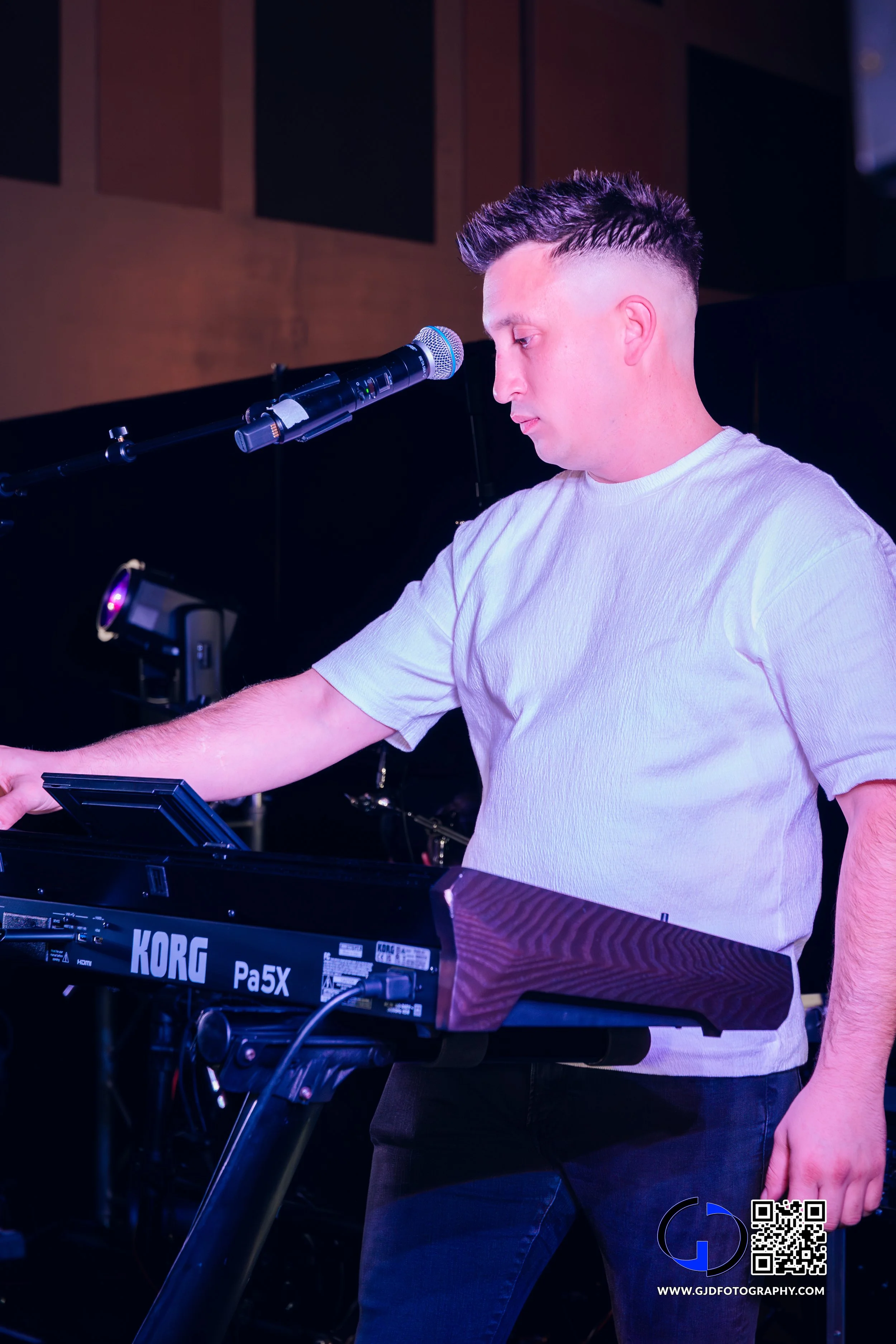 A young man wearing a white t-shirt playing a Korg keyboard on stage with a microphone positioned in front of him.