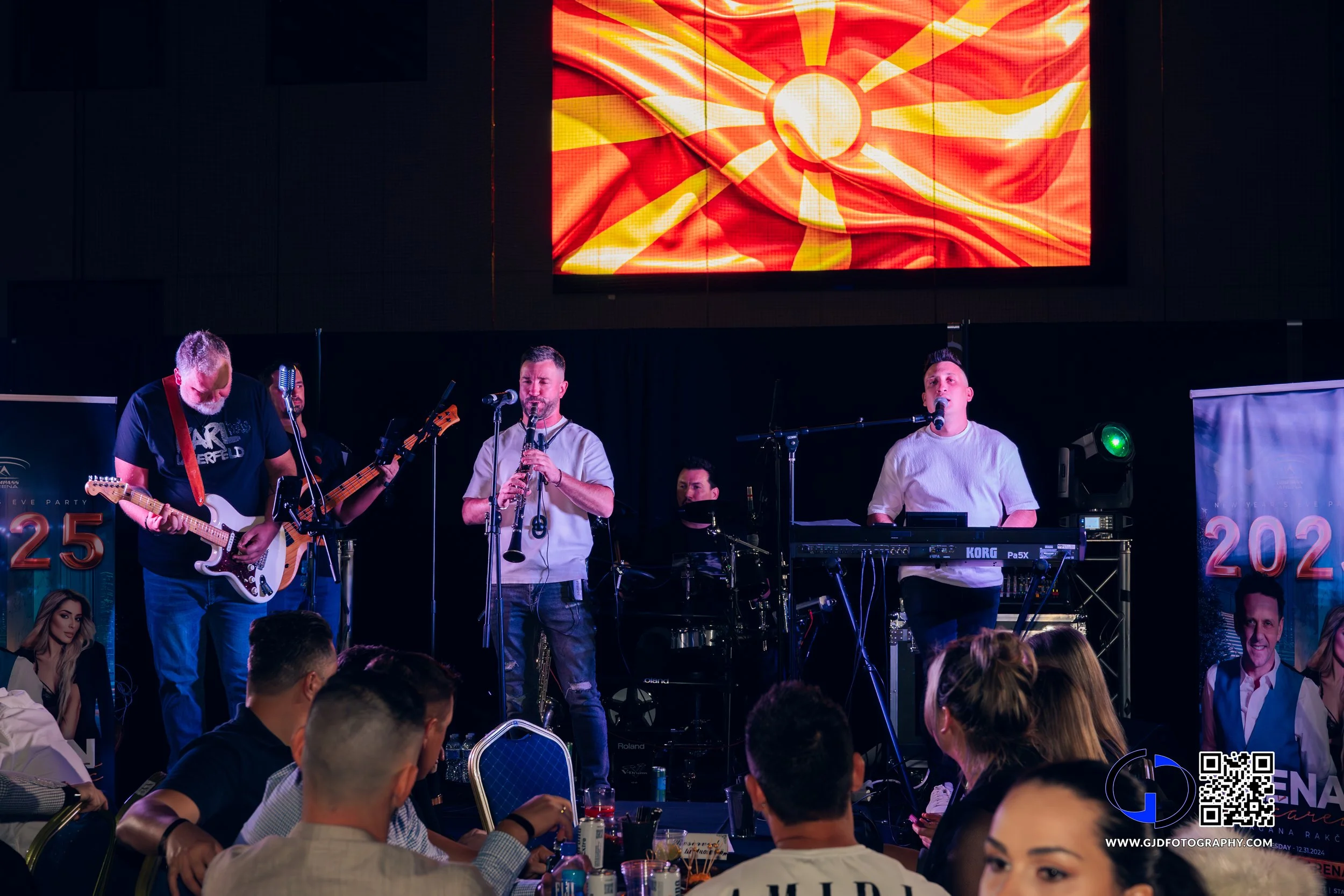 A live band performing on stage at an event, with a large screen displaying a stylized sun on the background, and an audience seated at tables watching the performance.
