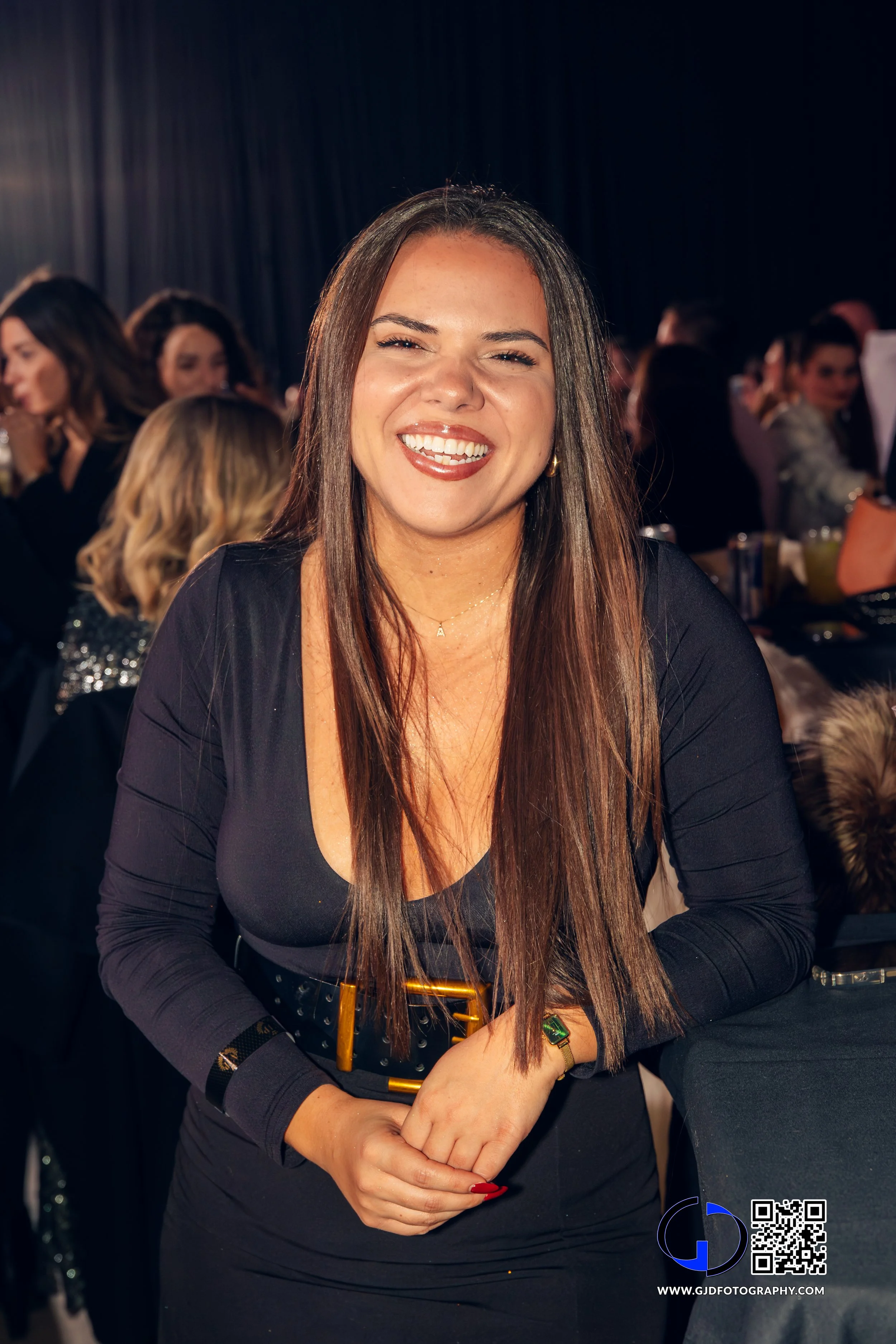 A smiling woman with long brown hair and a black dress standing at a crowded indoor event.
