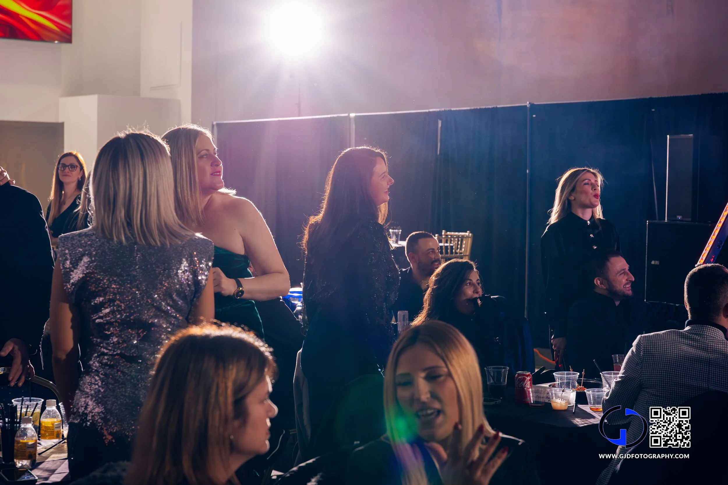 Group of people at a concert or event watching a performance with some standing and some seated, under dramatic lighting.