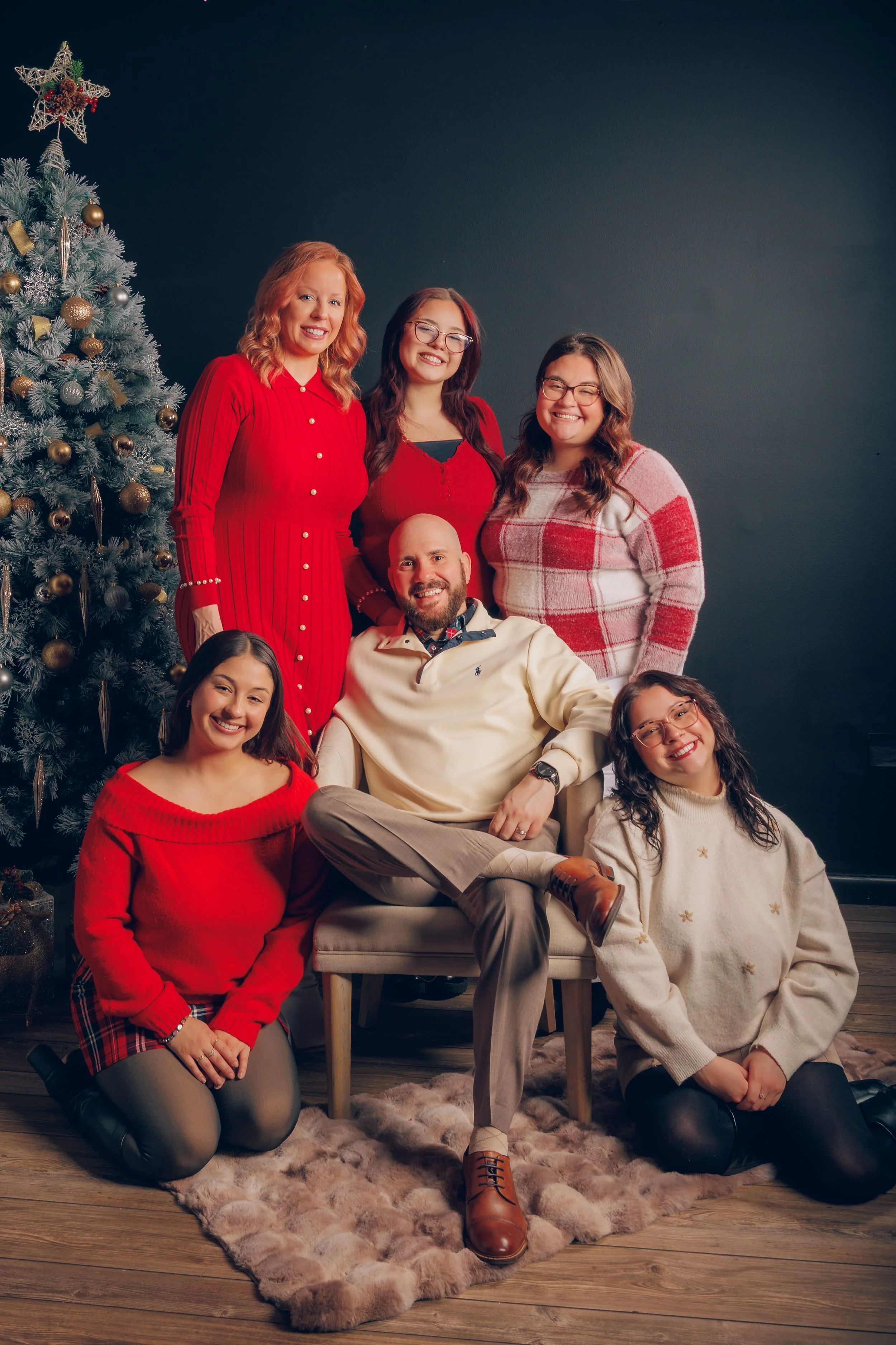 A group of seven people, including six women and one man, gathered around a Christmas tree decorated with gold and silver ornaments, posing indoors with a dark wall background. They are all smiling, wearing festive clothing in red, white, and plaid p