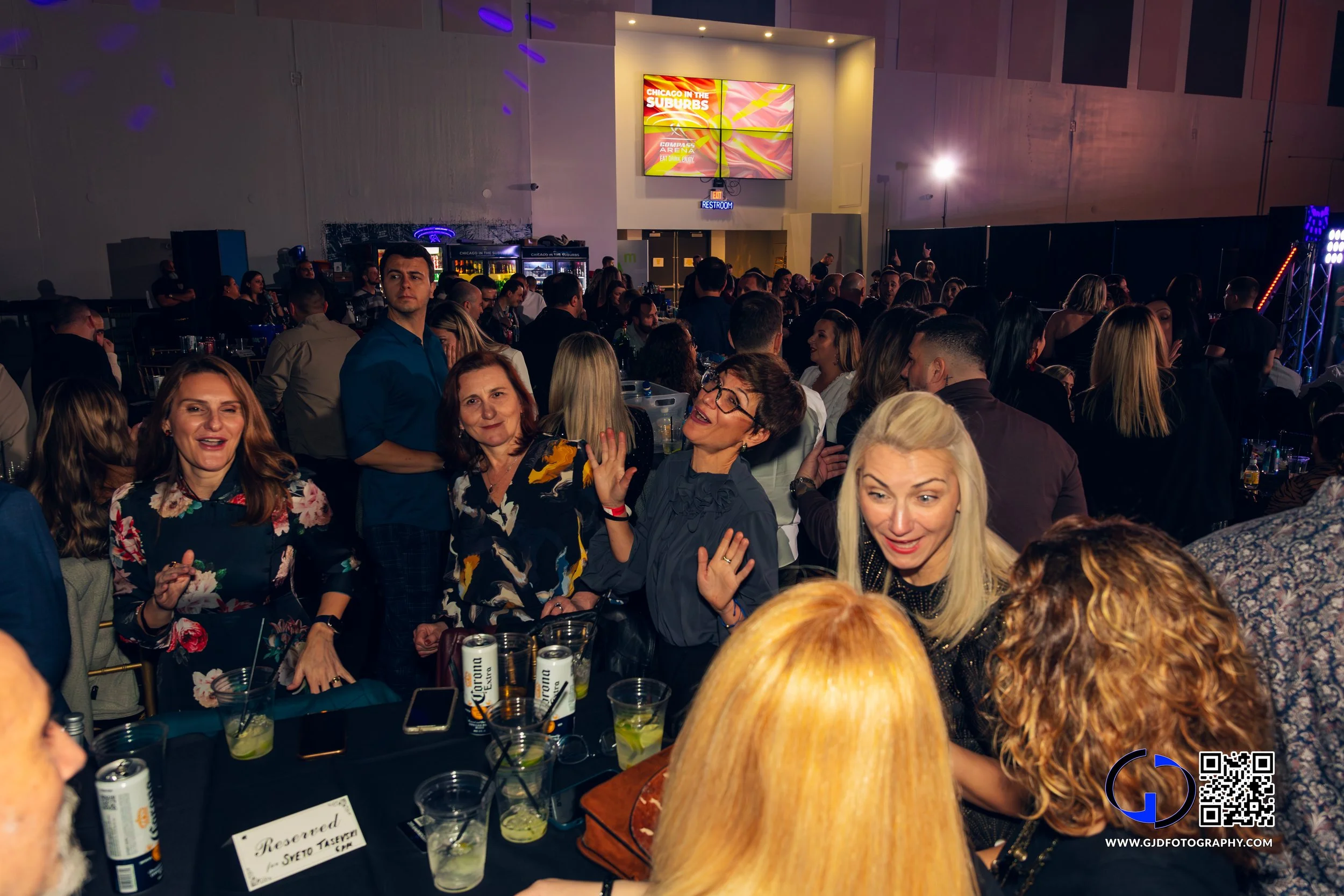 Crowd of people at an indoor event, some are seated at tables with drinks, others are standing and socializing, in a dimly lit venue with a large screen displaying 'Chicago in the Suburbs' in the background.