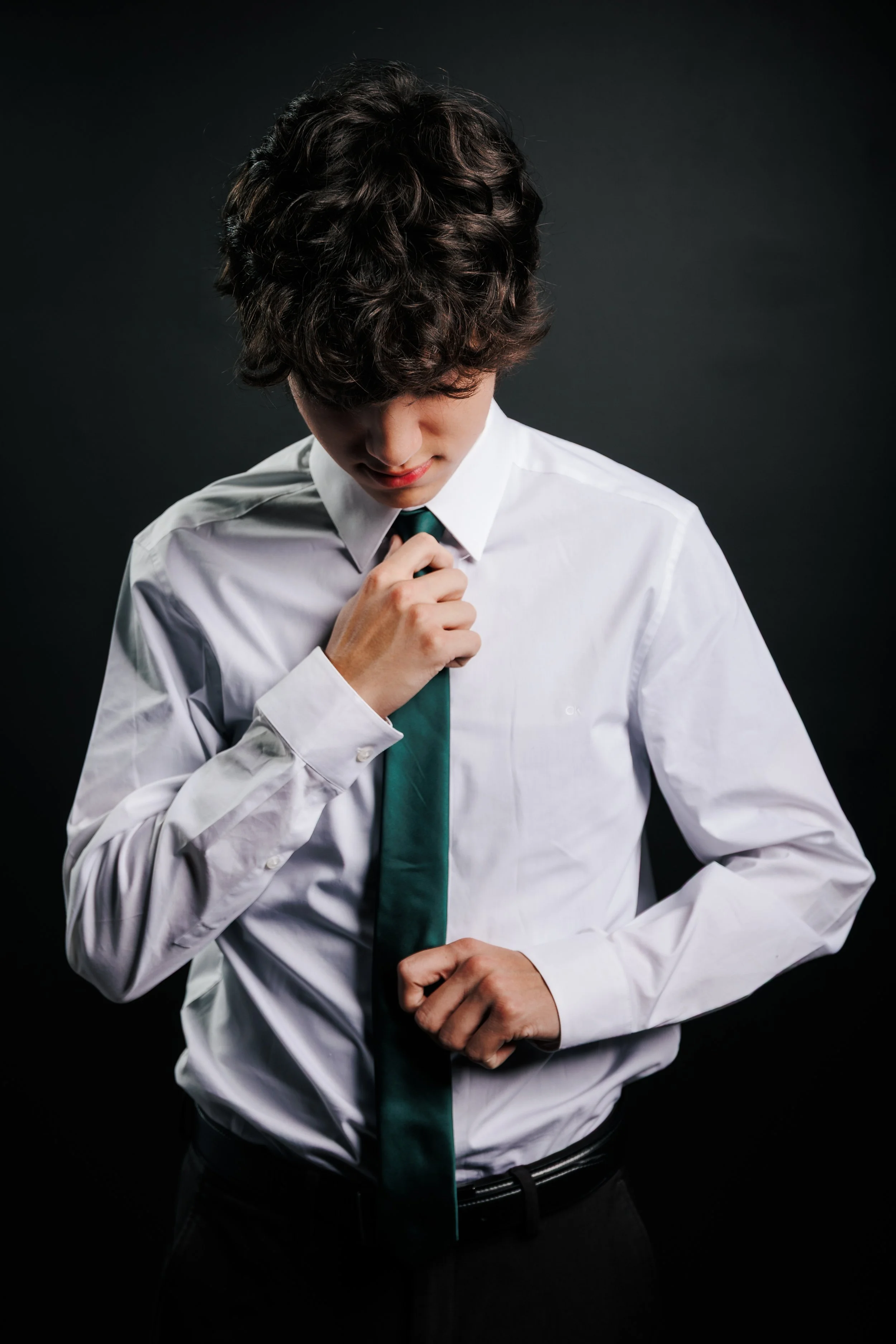 Young man with curly hair adjusting his green tie while wearing a white dress shirt against a dark background.