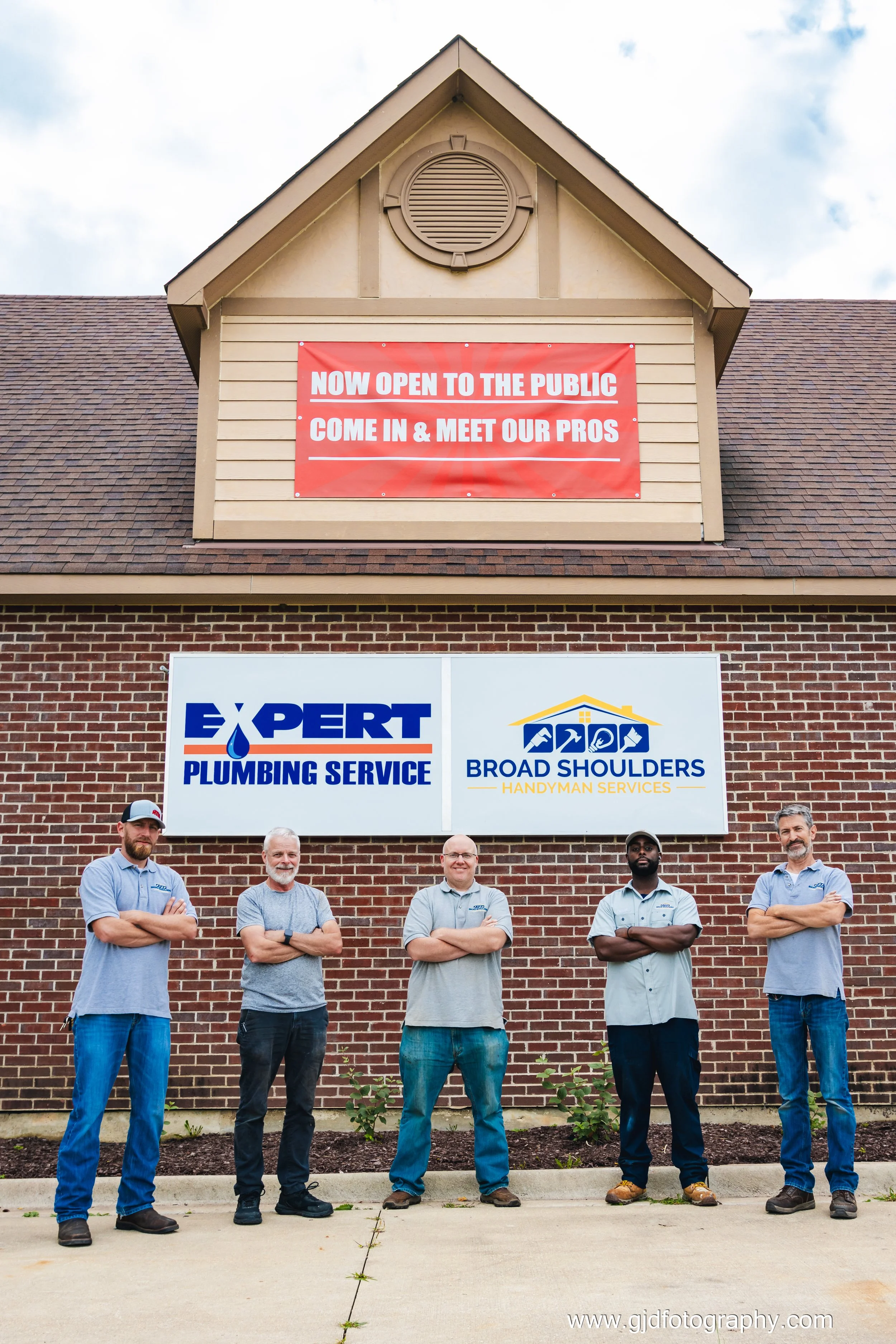Group of five men standing in front of a sign for Expert Plumbing Service and Broad Shoulders Handyman Services, with a banner above that reads 'Now open to the public, come in & meet our pros'.
