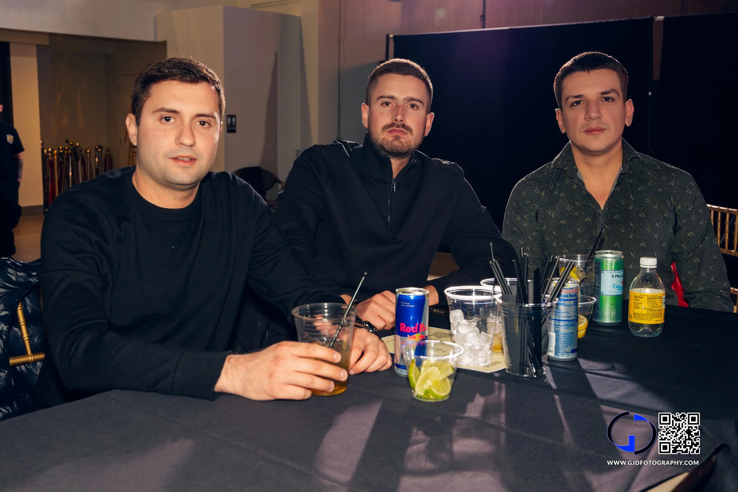 Three men sitting at a table with drinks and drink ingredients, in an indoor setting with dim lighting.