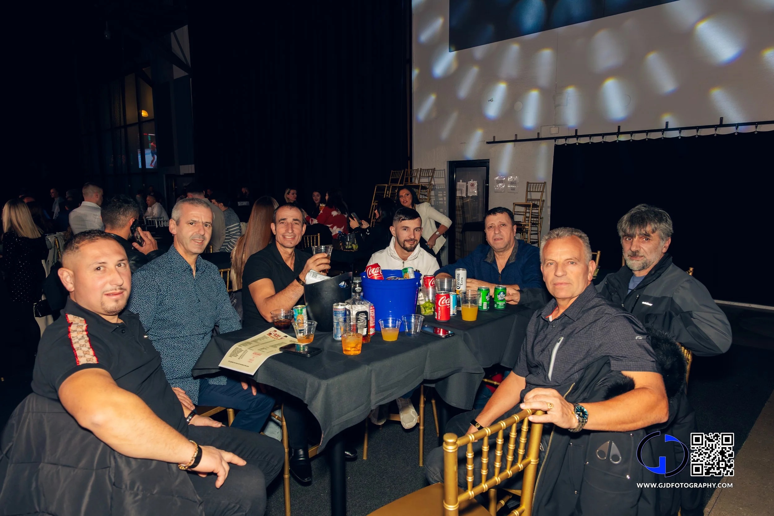 Group of seven men sitting around a banquet table at a social event, with drinks and snacks, in a dimly lit indoor venue.