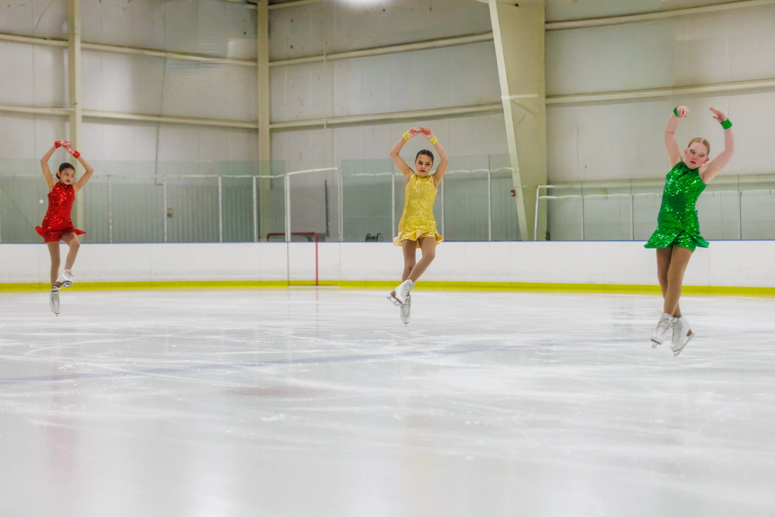 Three figure skaters in colorful costumes performing jumps on an ice rink inside an indoor ice skating rink.