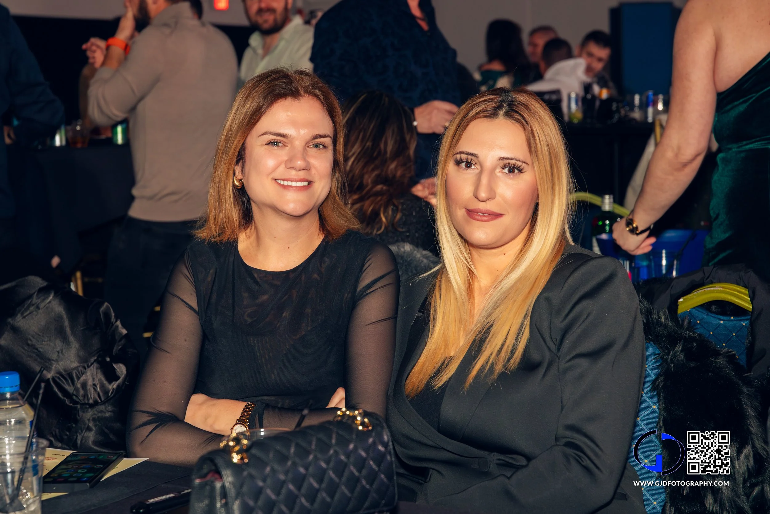 Two women sitting at a table during an event, smiling at the camera, with a crowded indoor background. One woman has shoulder-length brown hair and the other long blonde hair.