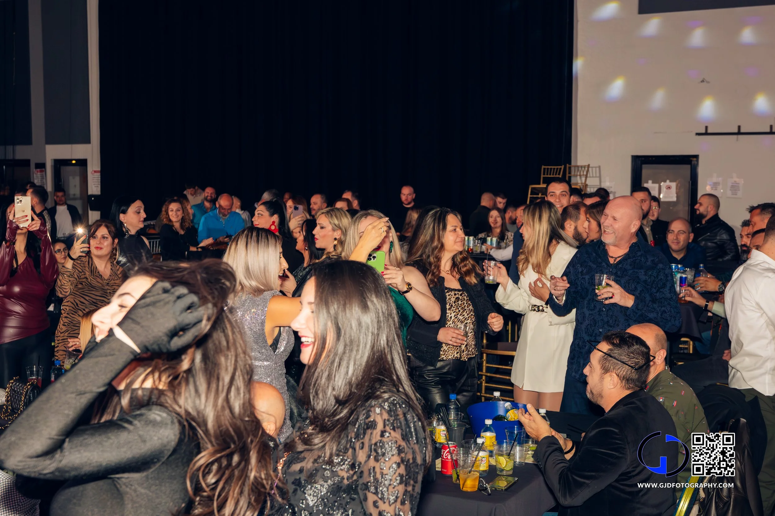 A lively crowd of people at a party or event, socializing, taking photos, and enjoying drinks in a large indoor venue with dark curtains and colorful lights.