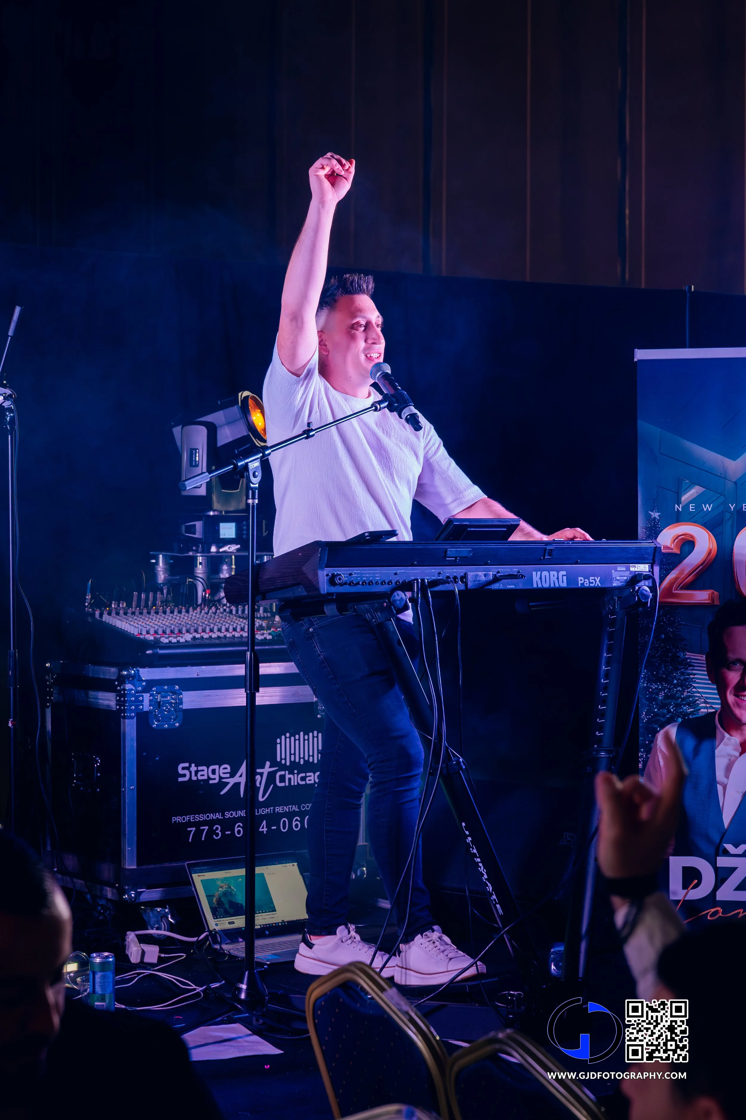 A man performing on stage, playing a keyboard and singing into a microphone, with his right arm raised in a fist, during a live music event.