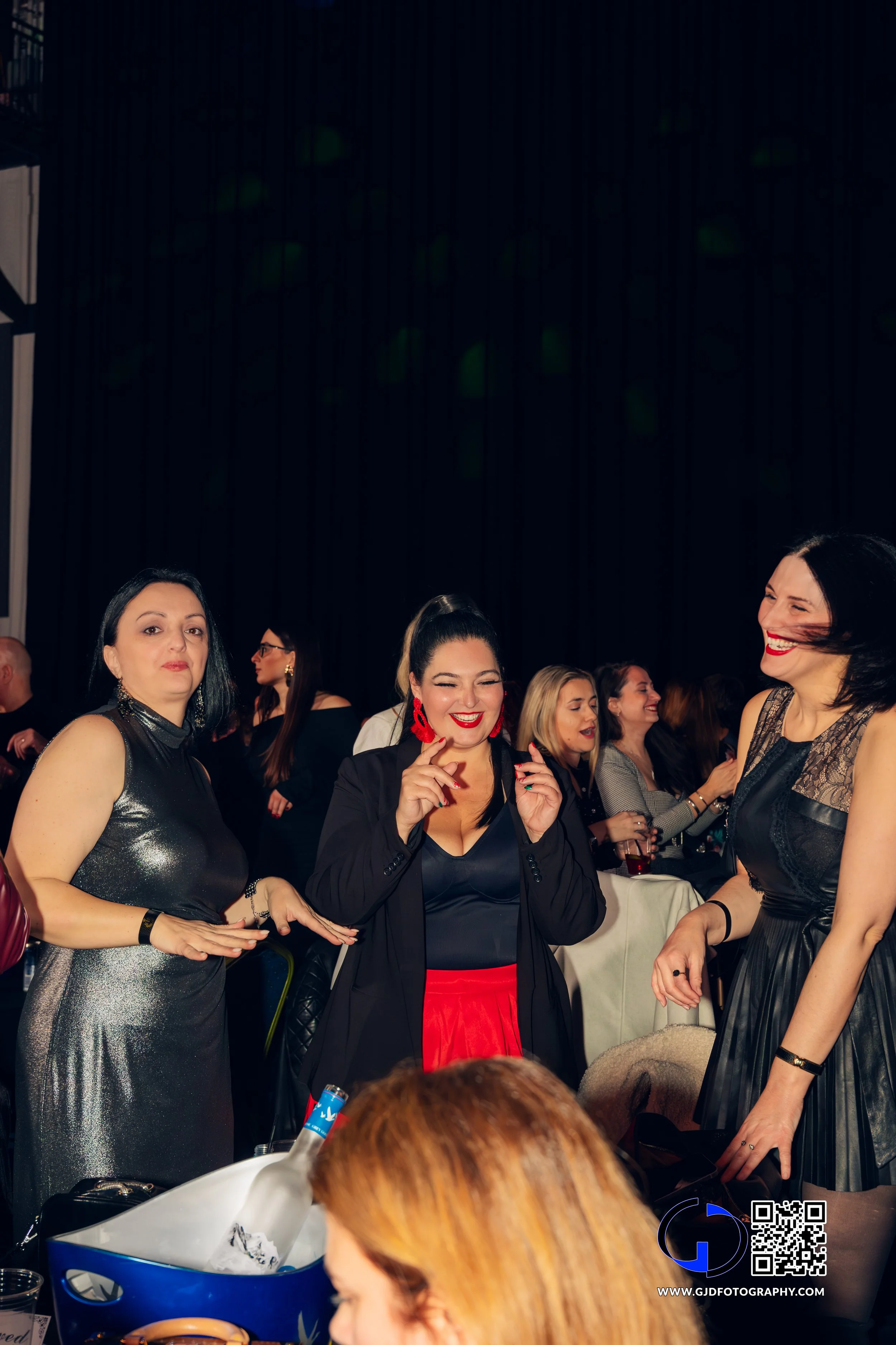 Women at a lively social event, smiling and talking with each other, dressed in elegant evening attire.