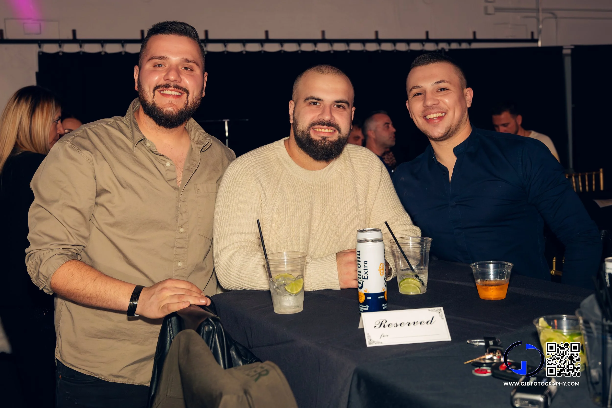 Three young men are sitting at a table, smiling, with drinks in front of them, at an indoor social event. One man is standing, and there are other people in the background.