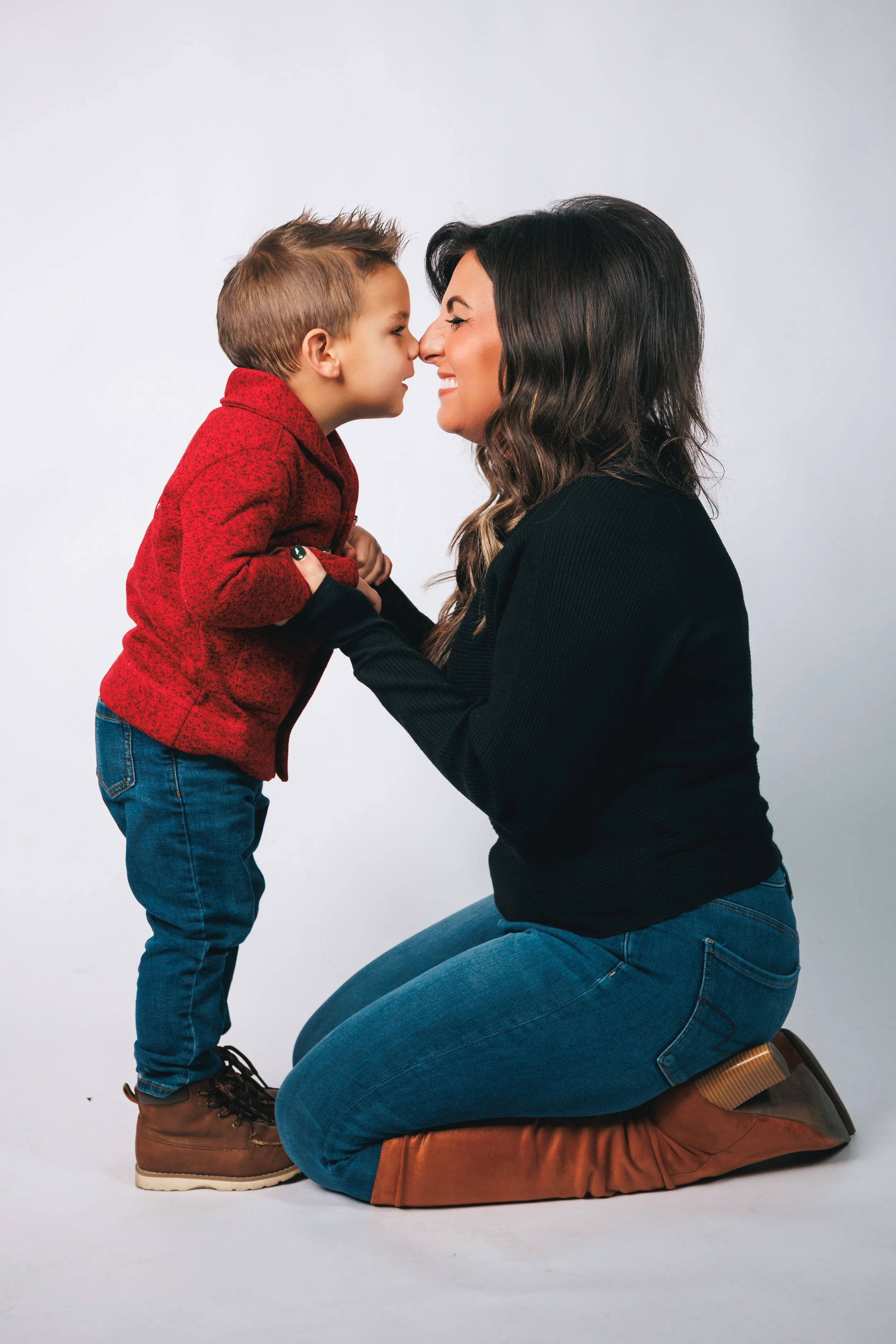 A woman kneeling and smiling at a young boy with their faces close, noses touching, against a plain white background.