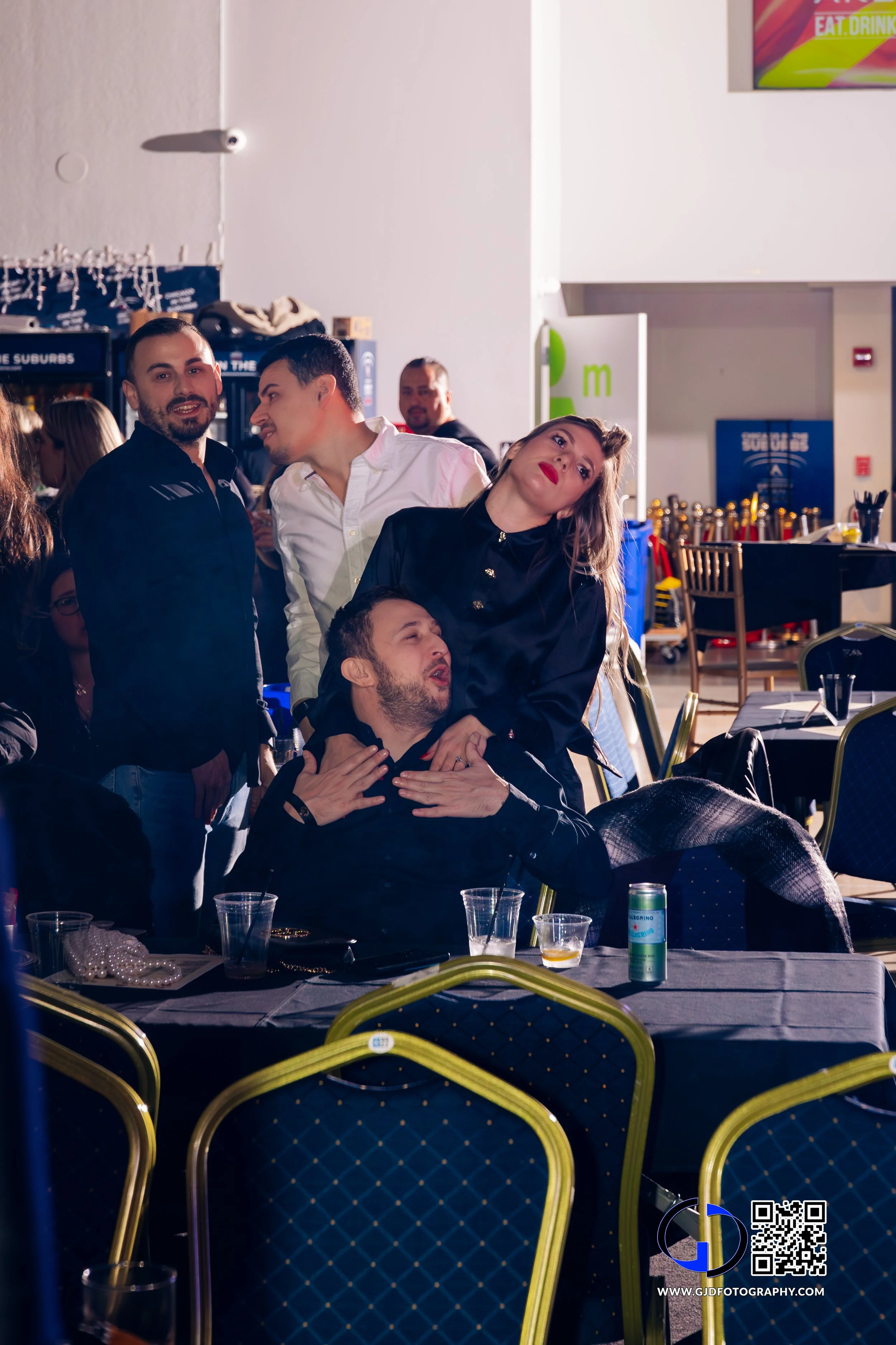 Group of five people at a social event, some sitting at tables and some standing behind them, with one person appearing as if being playfully pinched by another. The setting has tables with drinks and tableware, and a brightly lit background with dec