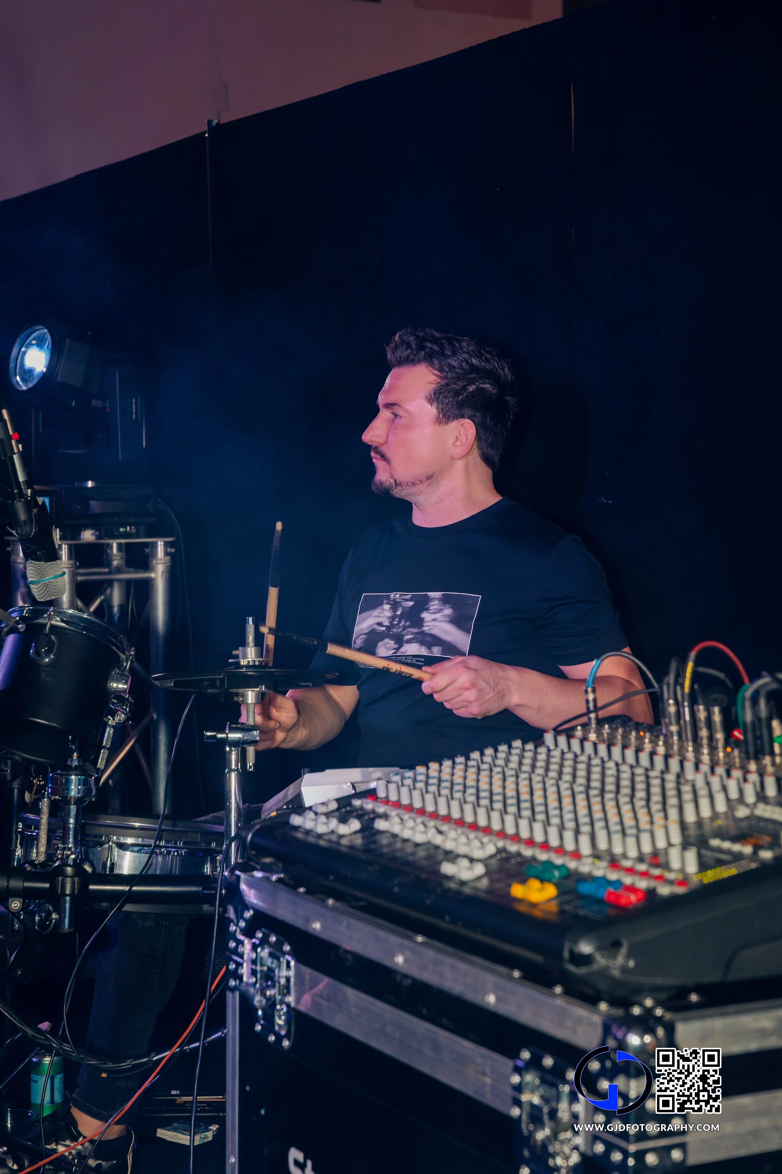 A male musician playing electronic drums during a live performance, with a sound mixer and stage lighting in the background.